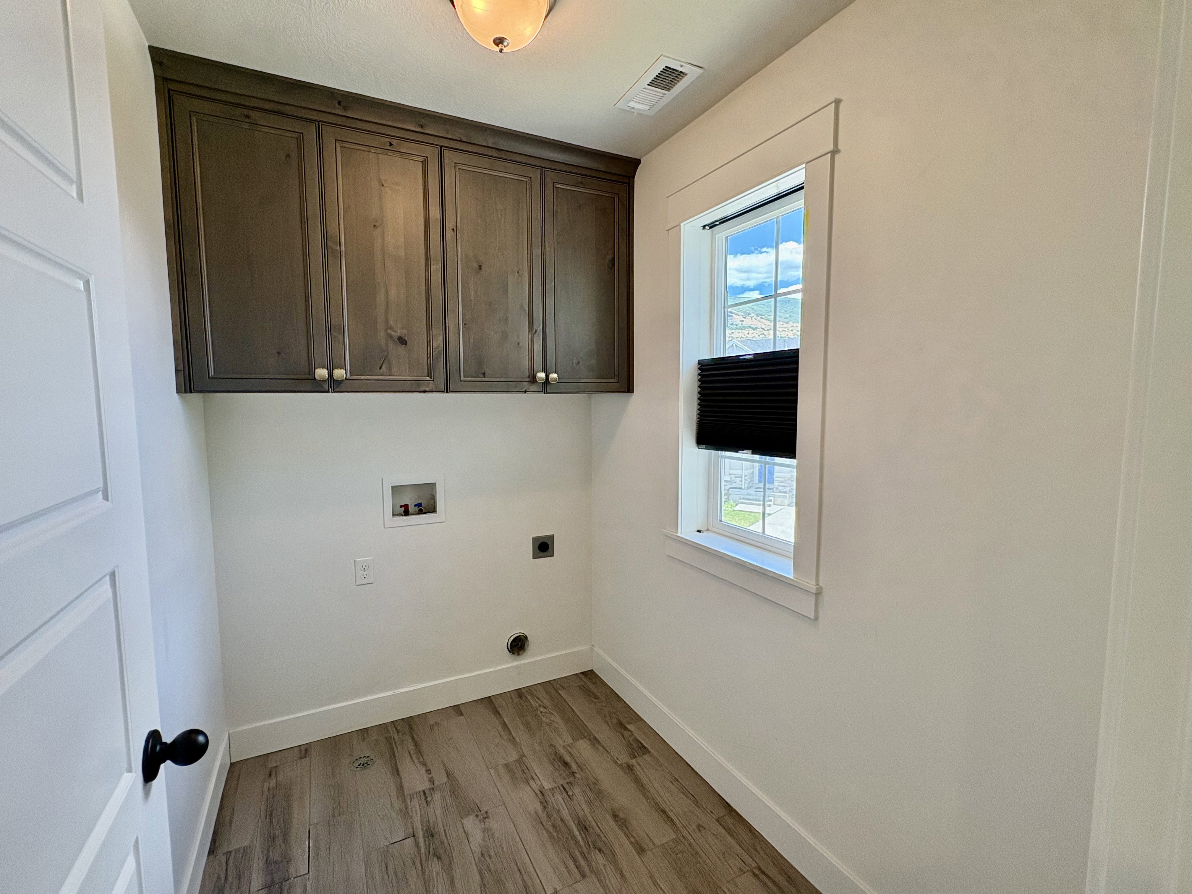 Laundry area featuring wood-finish flooring, overhead cabinetry with brass hardware, a large window, and white trim