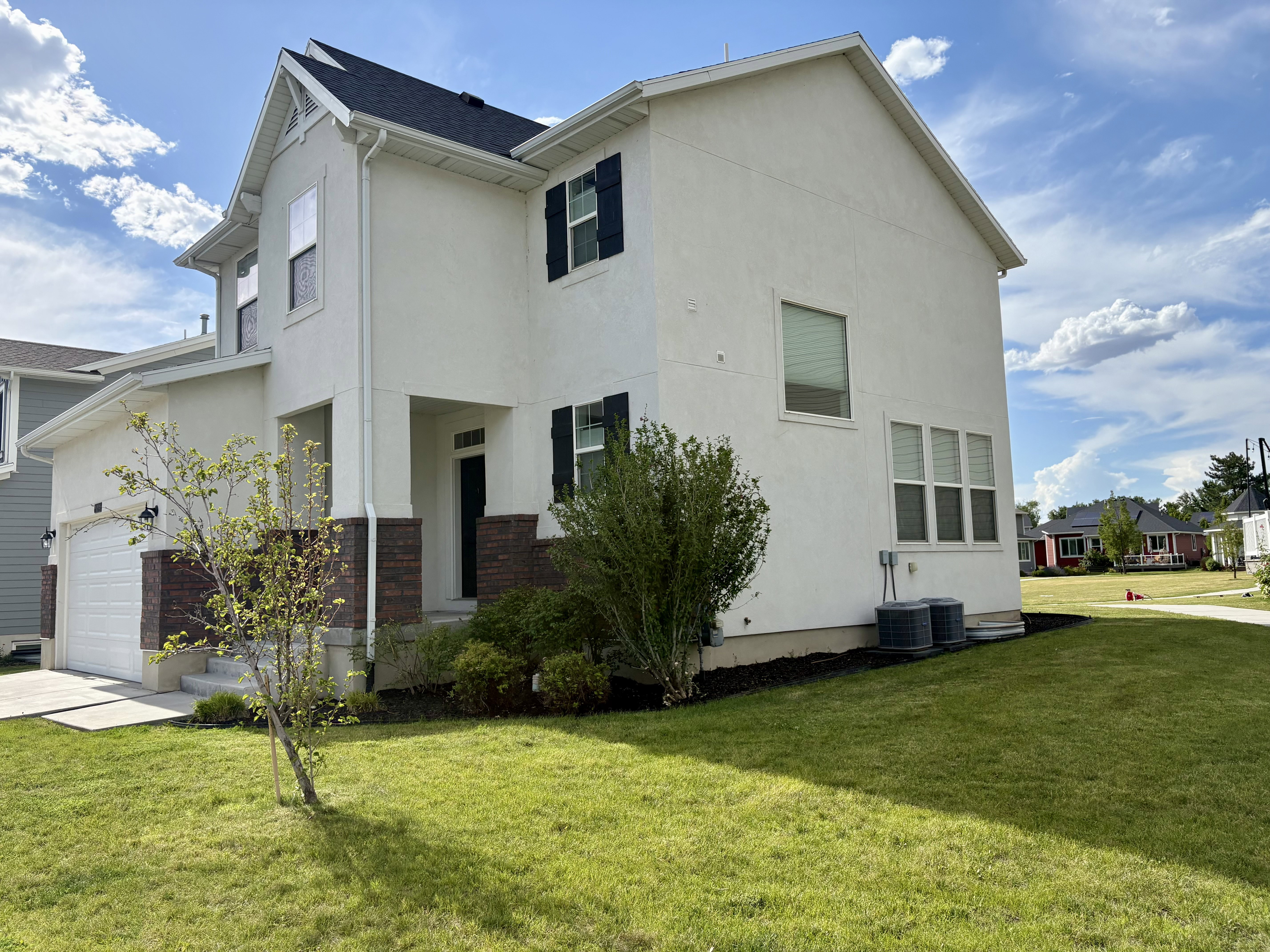 Two-story stucco residence featuring dark brick accents, black window shutters, a front entry porch, an attached garage, and a lush green lawn