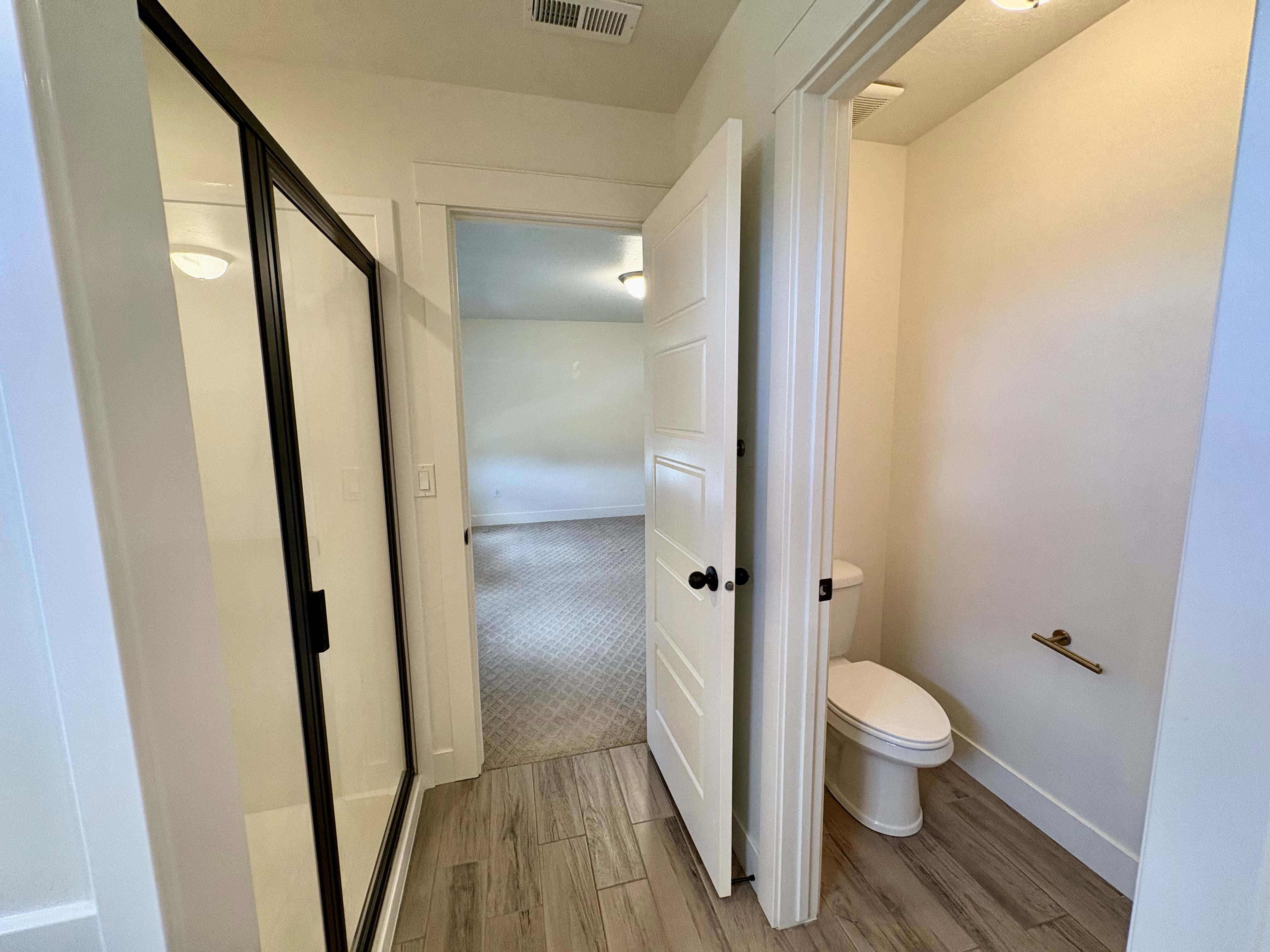 Bathroom featuring wood-finish flooring, a frameless shower with a black trim, a paneled interior door with black hardware, and a separate toilet area with a brass-finish towel bar