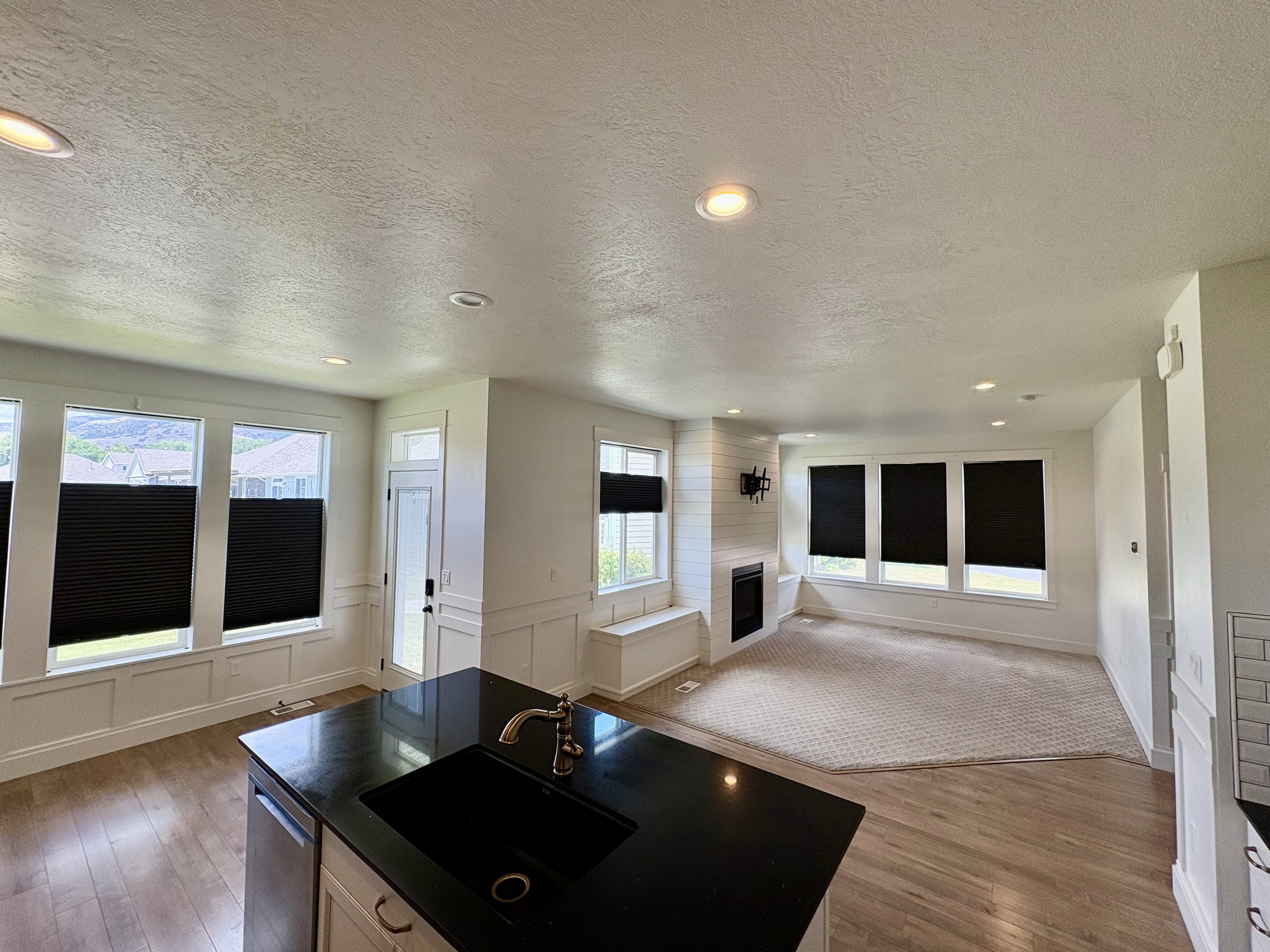 Open-concept interior featuring wood-finish flooring, a kitchen island with a black sink and dark countertop, a fireplace with a white shiplap surround, recessed lighting, and large windows with blinds