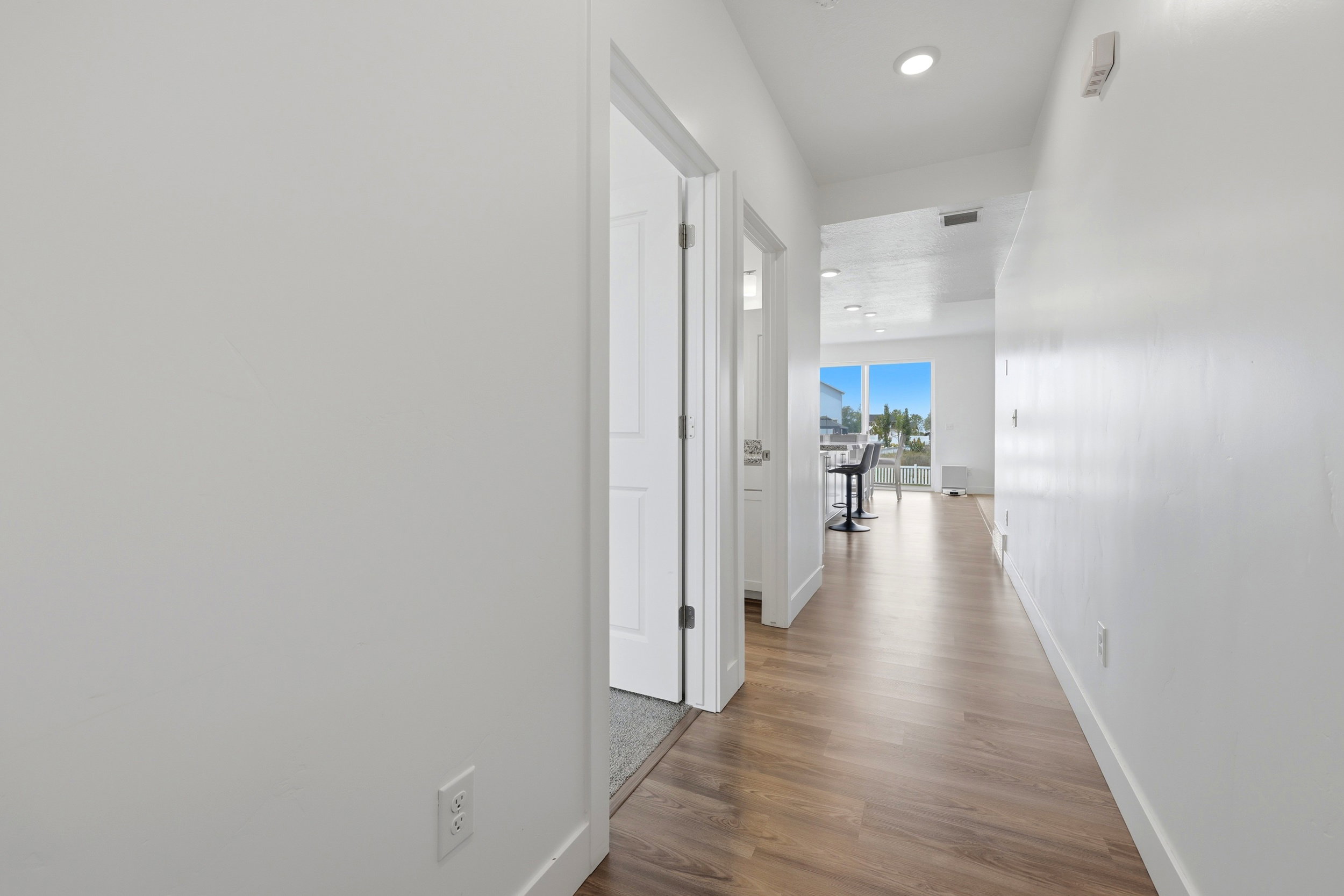 Hallway featuring wood-finish flooring, white walls, recessed lighting, and a sightline to a bright living area with large windows