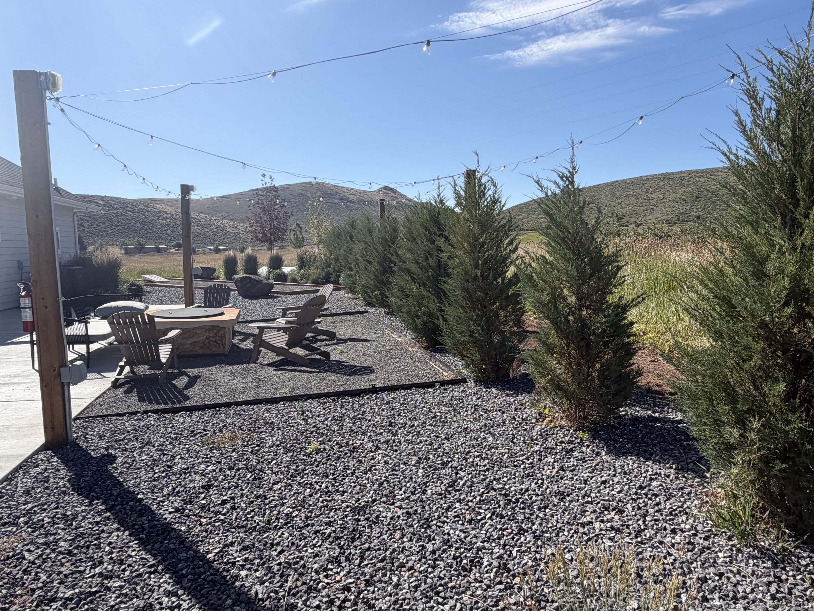 Outdoor living area featuring gravel landscaping, a built-in fire pit, string lighting, and mature evergreen privacy plantings