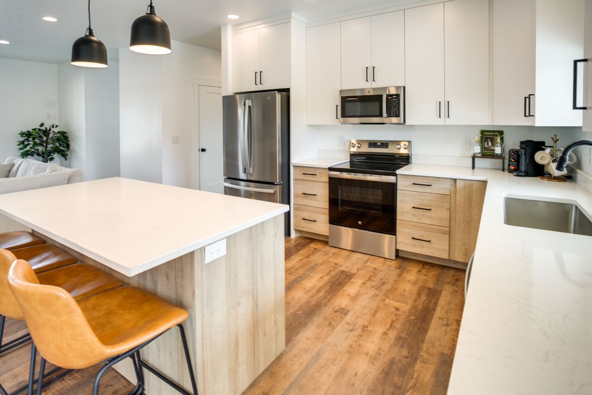 Modern kitchen featuring a large island with a light-toned countertop, stainless steel appliances, light wood-finish flooring, and two-toned cabinetry with dark hardware