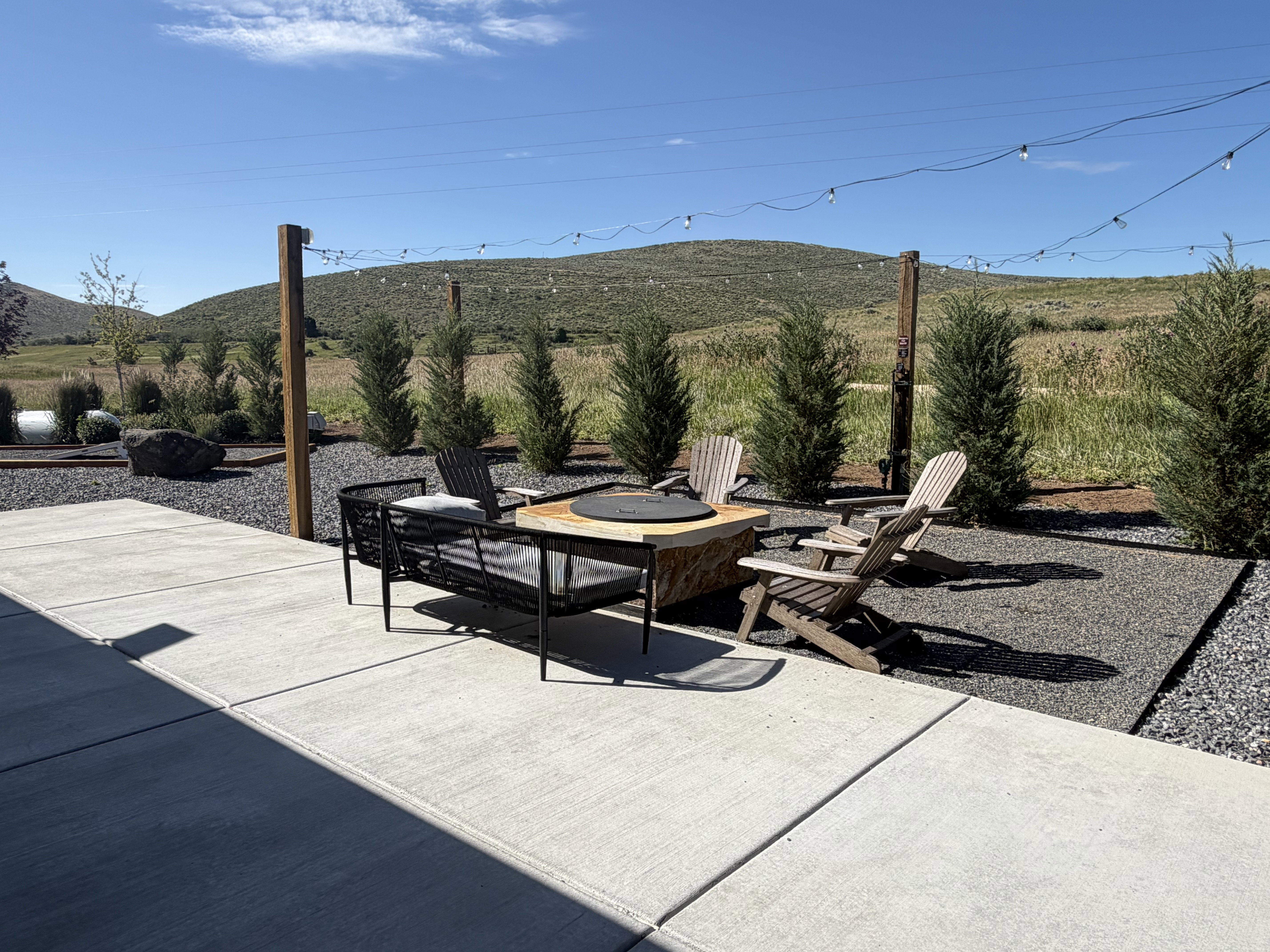 Expansive concrete patio area featuring a dedicated gravel fire pit zone with string lighting suspended from wooden posts