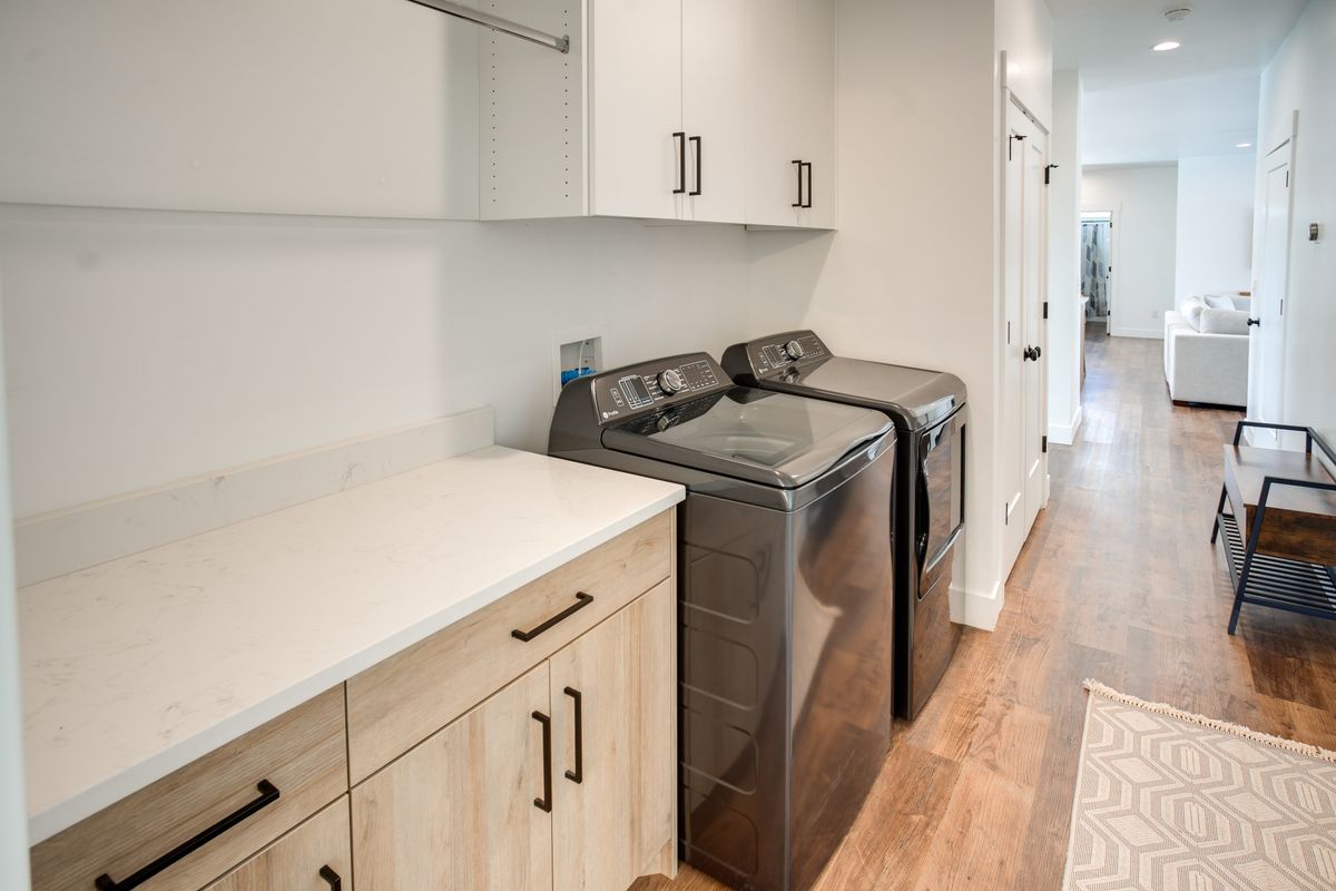 Laundry area featuring wood-finish flooring, light-toned base cabinetry with dark hardware, white upper cabinetry, a light-colored countertop, and a built-in hanging rod