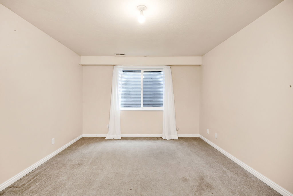 Second basement bedroom at the end of the hall featuring neutral carpeting, light-toned walls, and a single window with horizontal blinds