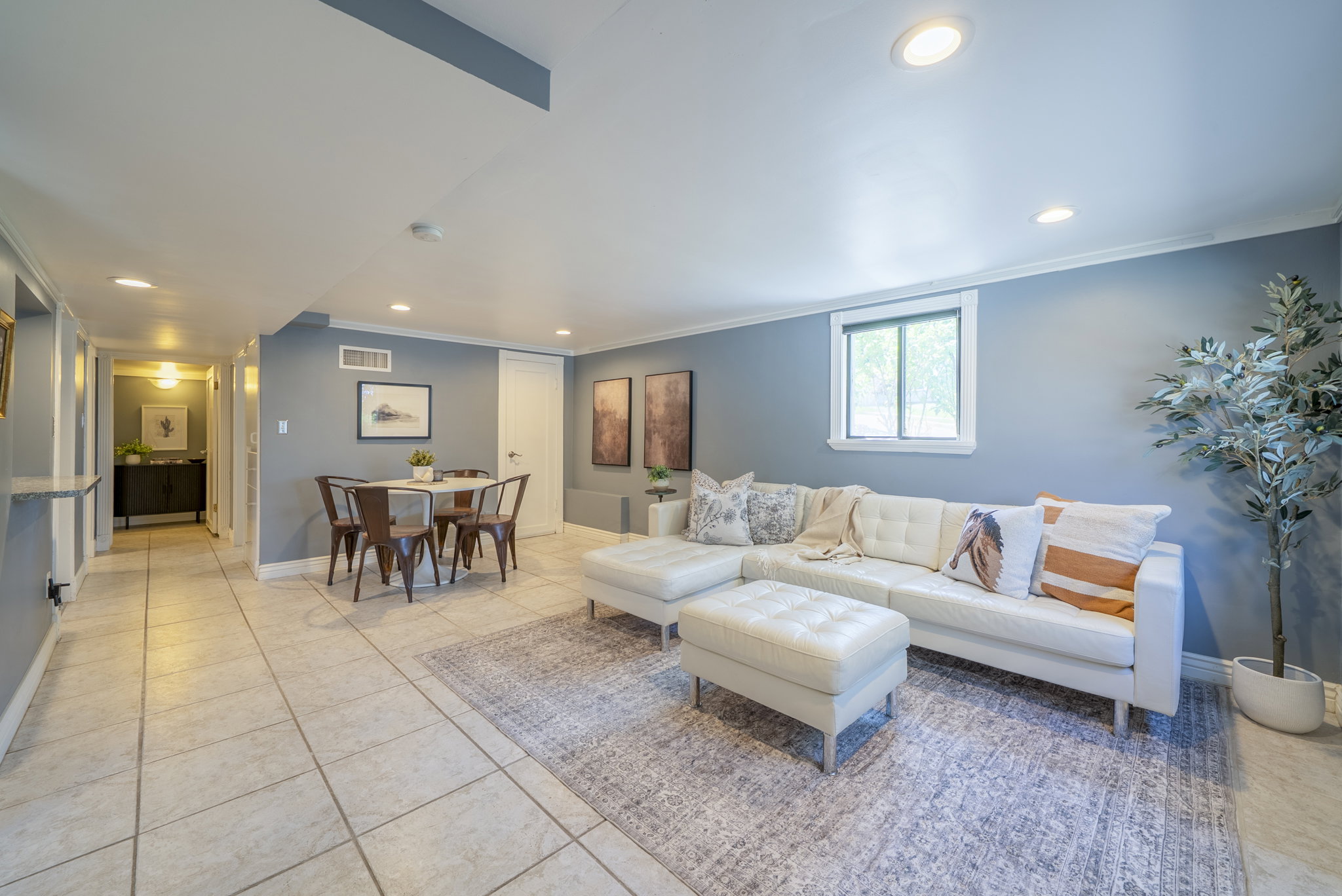 Spacious open-concept room featuring light-toned tile flooring, recessed lighting, crown molding, and a window providing natural illumination
