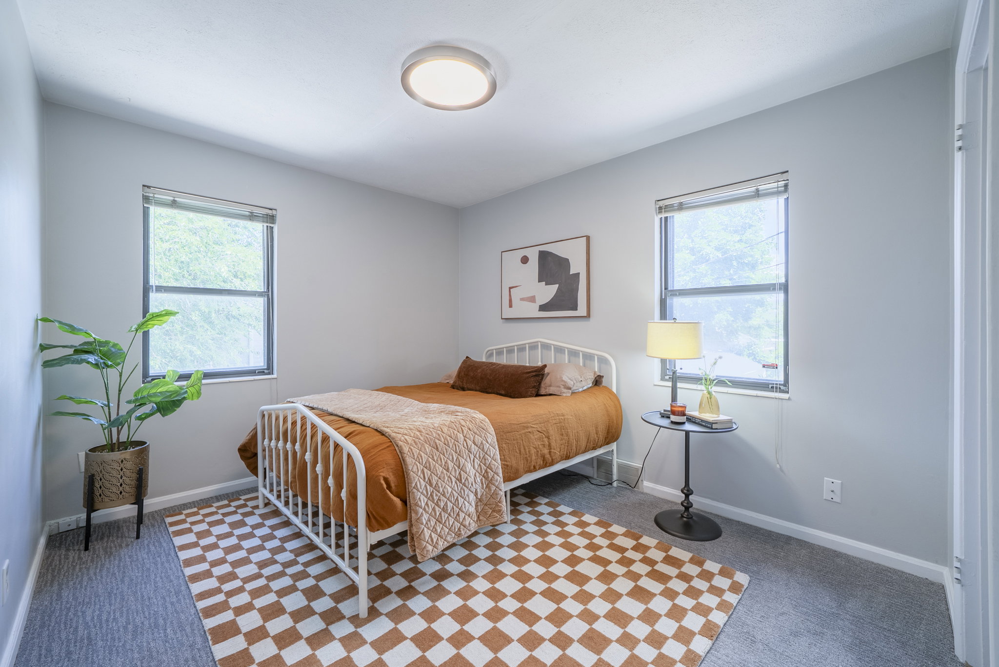 Neutral-toned room featuring a flush-mount ceiling light fixture, two windows with blinds, a checkerboard pattern area rug over carpeting, and baseboard trim