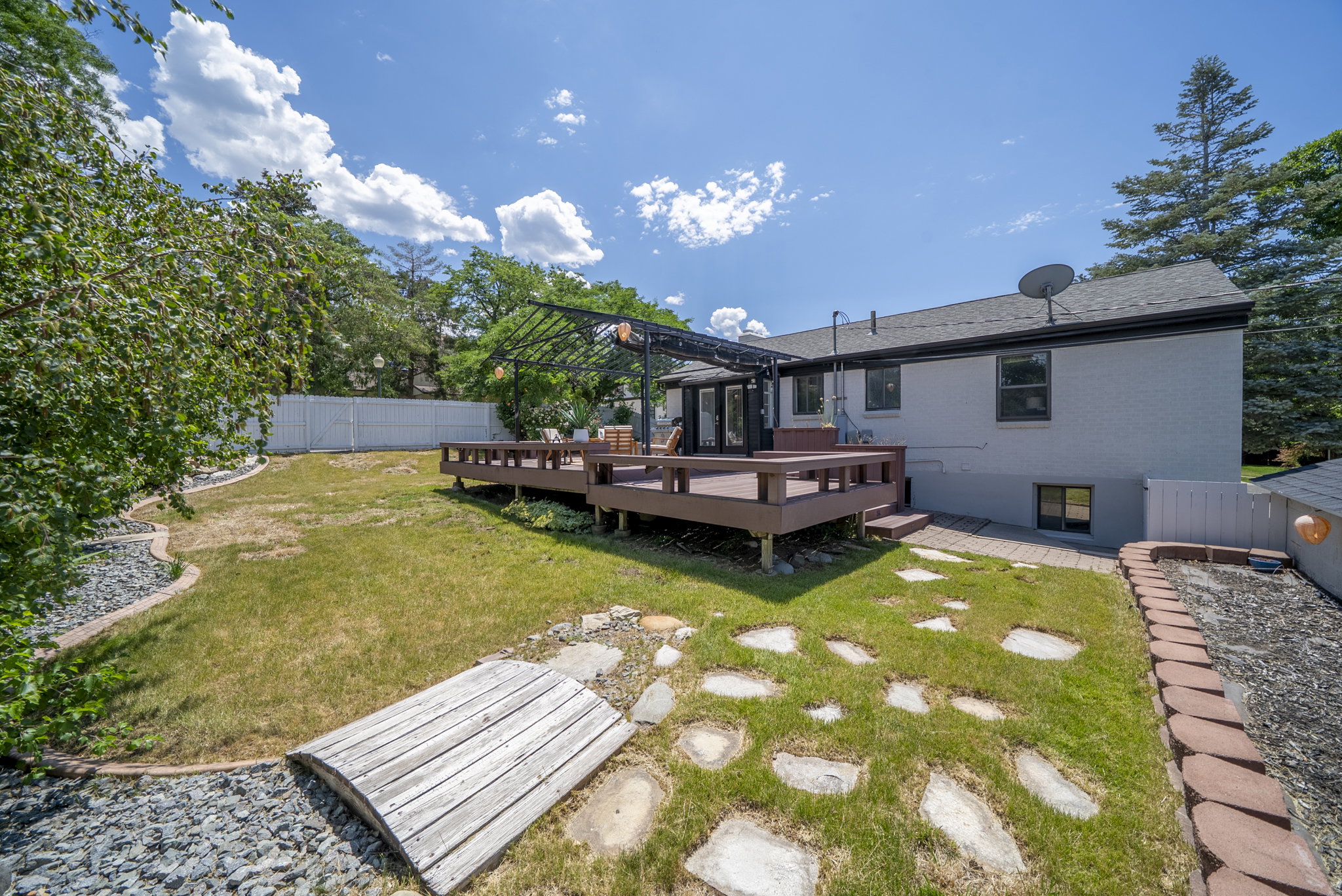 Expansive backyard featuring a multi-level wooden deck with a pergola, a manicured lawn, stone pathway, and a decorative wooden bridge over a rock bed