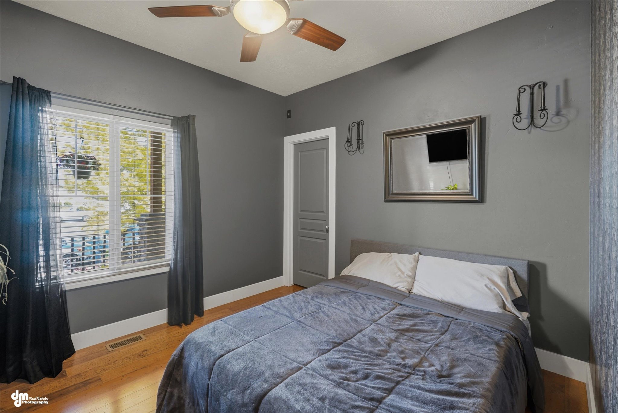 Spacious room featuring wood-finish flooring, a ceiling fan with integrated lighting, a large window with blinds, and a paneled door