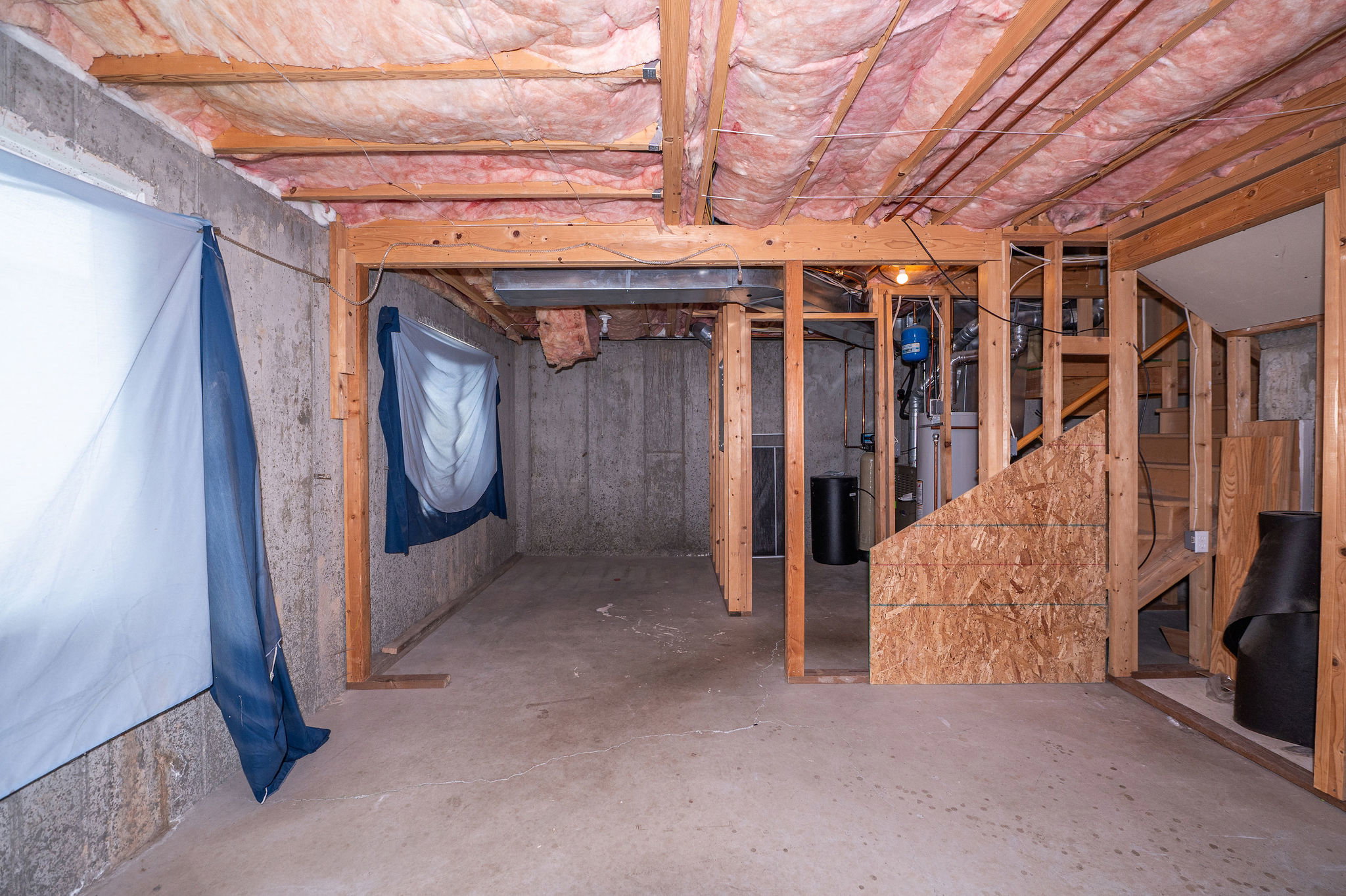 Unfinished basement featuring concrete walls and flooring, exposed wood framing, ceiling insulation, and a partial staircase