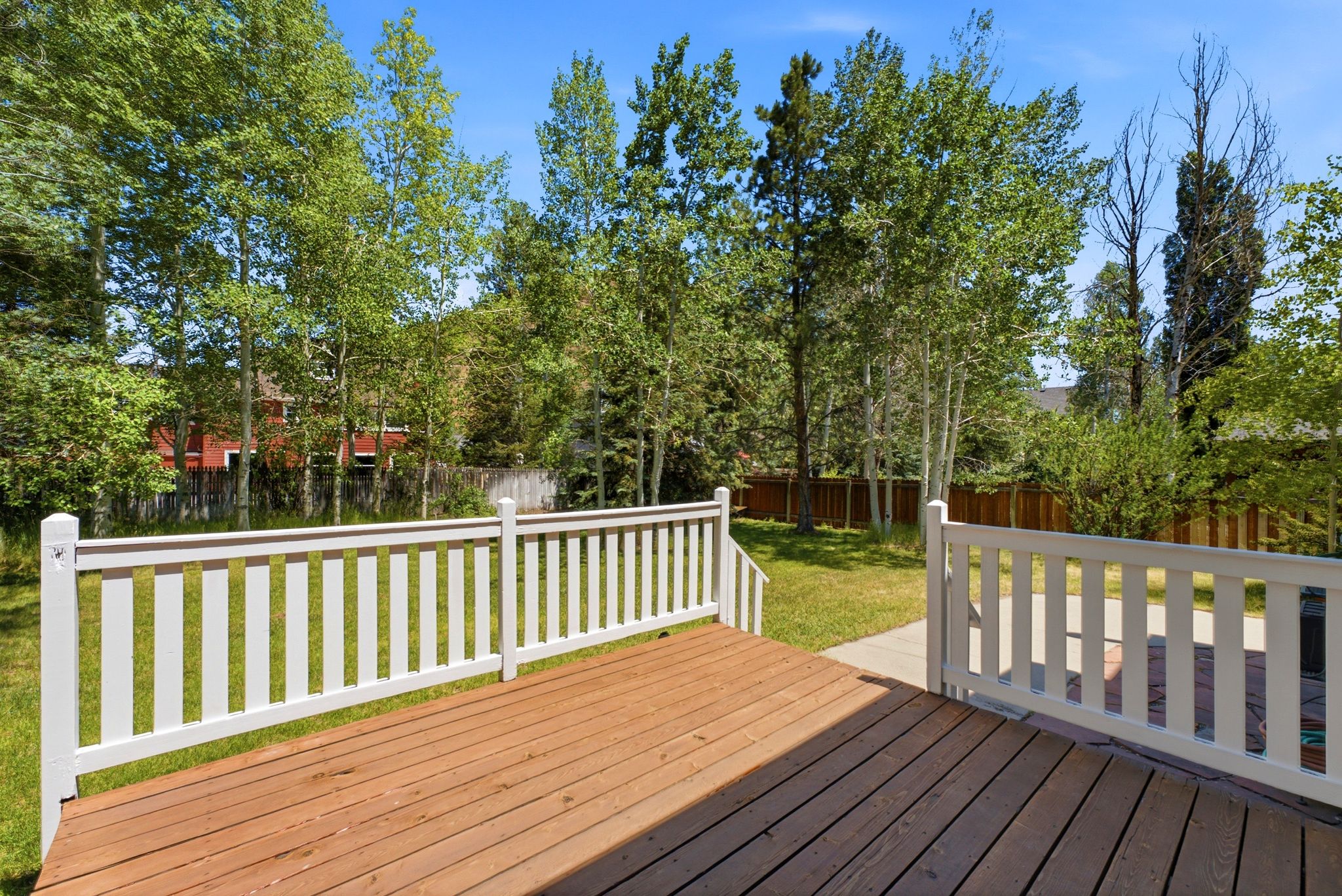 Wood-finish deck with white railing, featuring a verdant lawn, mature trees, and a concrete pathway