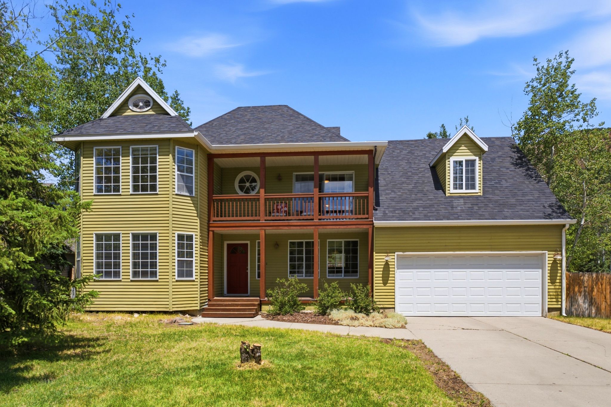 Green siding exterior featuring a two-car garage, upper-level balcony, dormer window, and a prominent front entrance with a red door