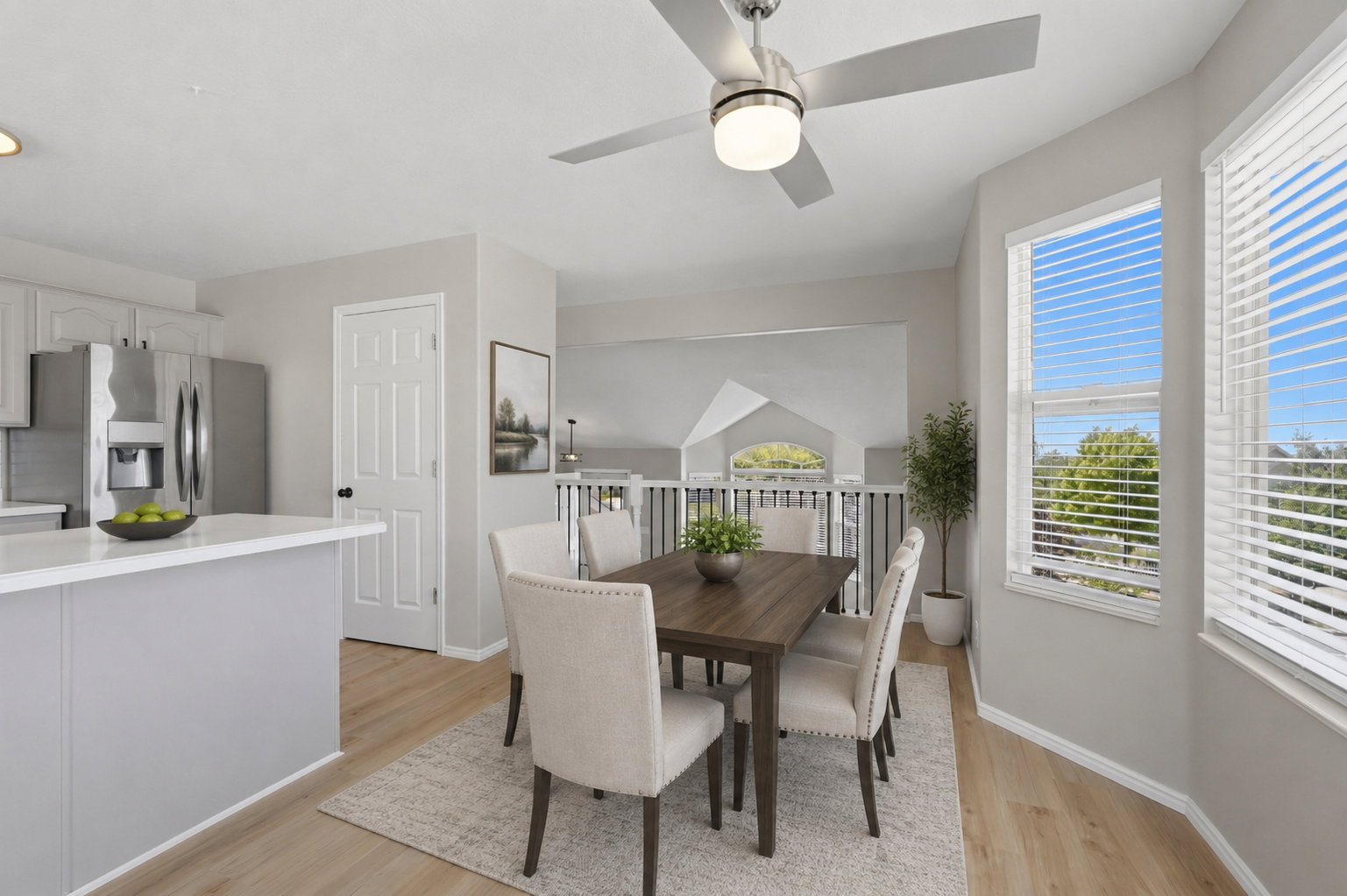 Open-concept dining area featuring wood-finish flooring, a contemporary ceiling fan with integrated lighting, and multiple windows with white blinds. Staged virtually.