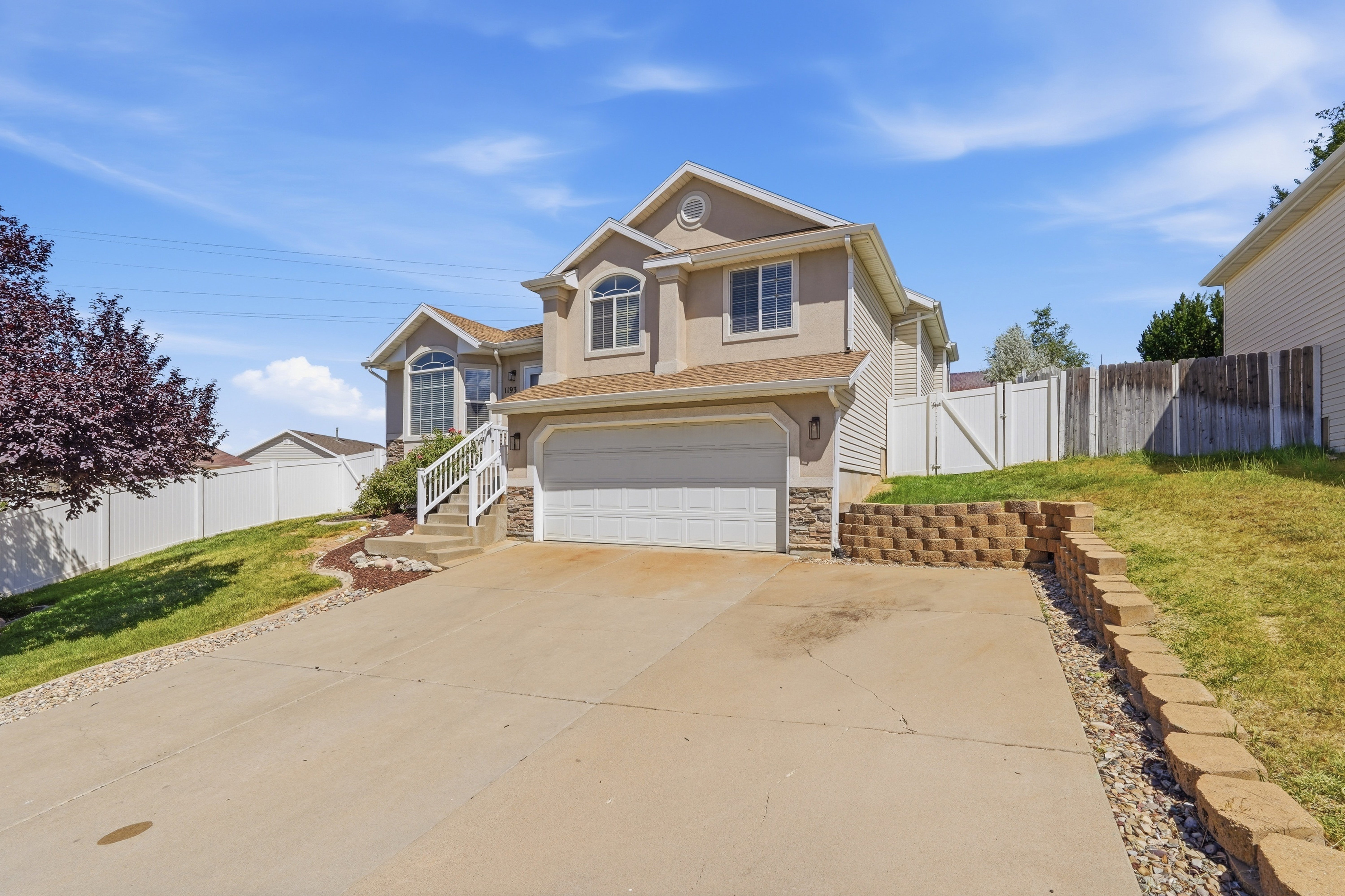 Two-story stucco and siding residence featuring a two-car garage, stone accent siding, concrete driveway, and a retaining wall