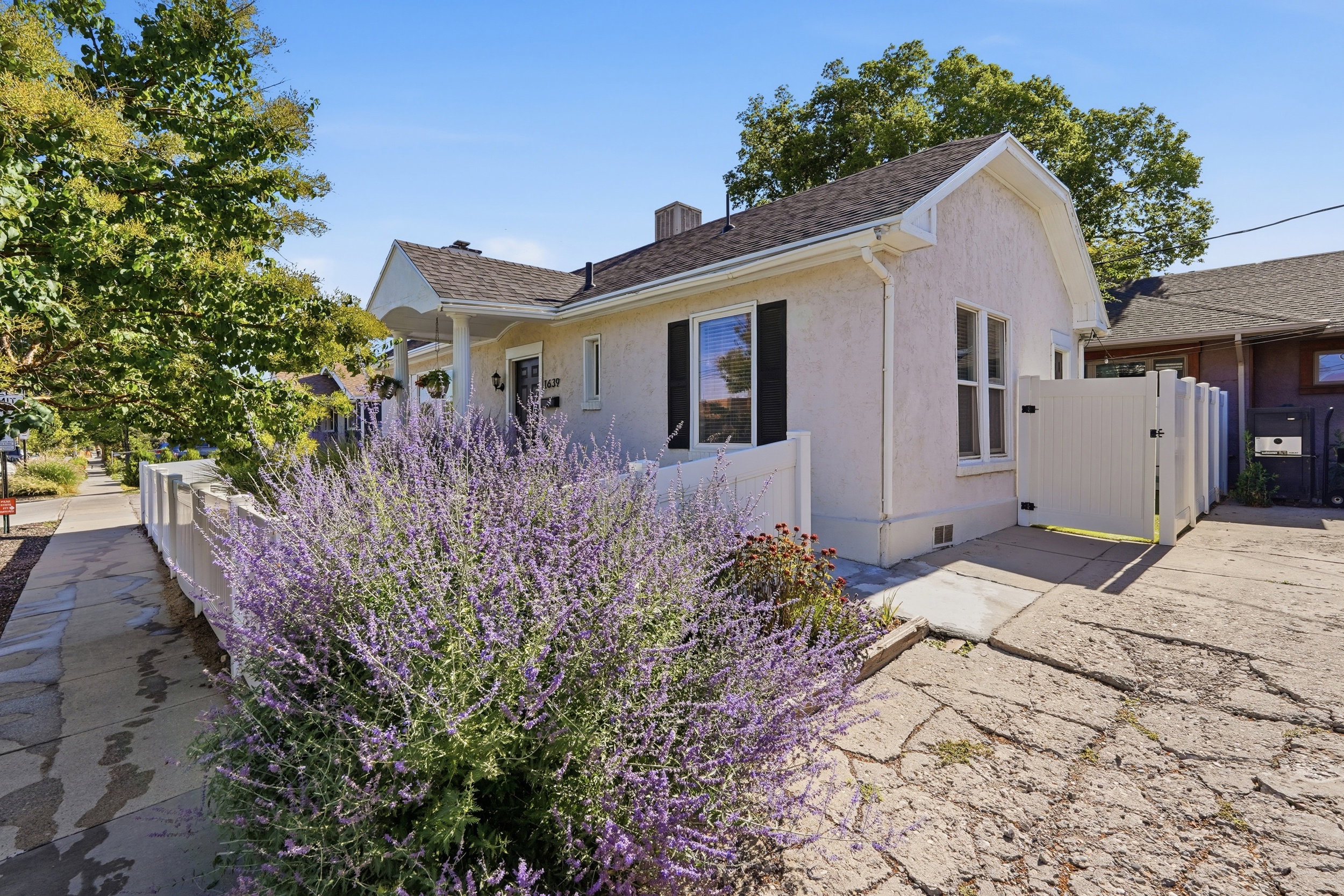 Stucco exterior home featuring a covered front porch, white picket fence, mature landscaping, dark shutters, and a private side gate