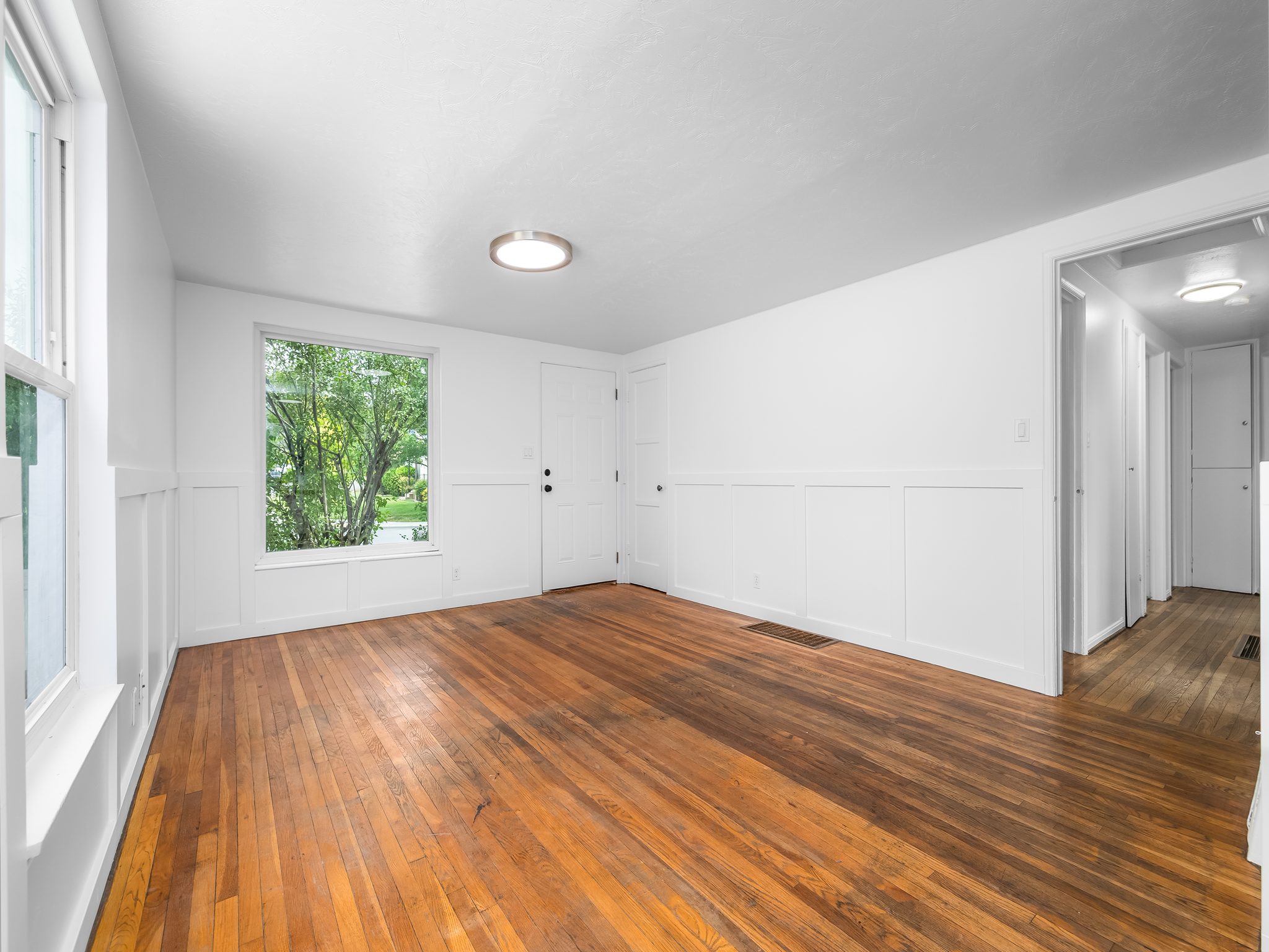 Spacious interior featuring wood-finish flooring, white paneled wainscoting, large window, and flush-mount ceiling light