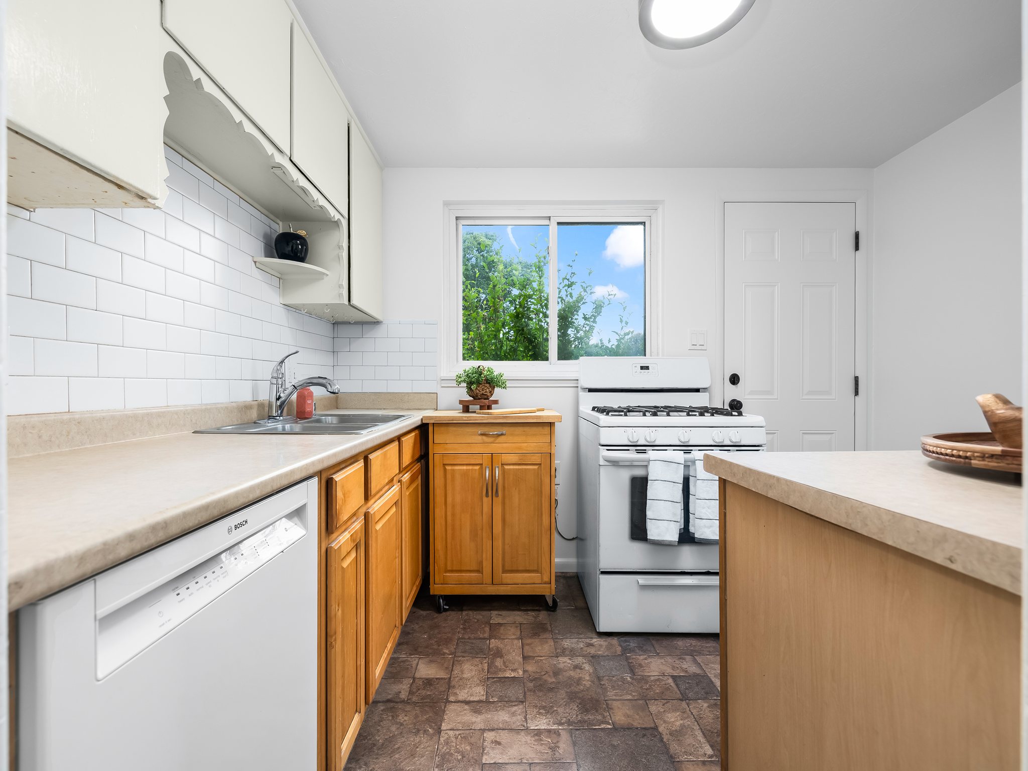 Kitchen featuring a double basin stainless steel sink, white subway tile backsplash, light wood-finish cabinetry, and neutral countertops