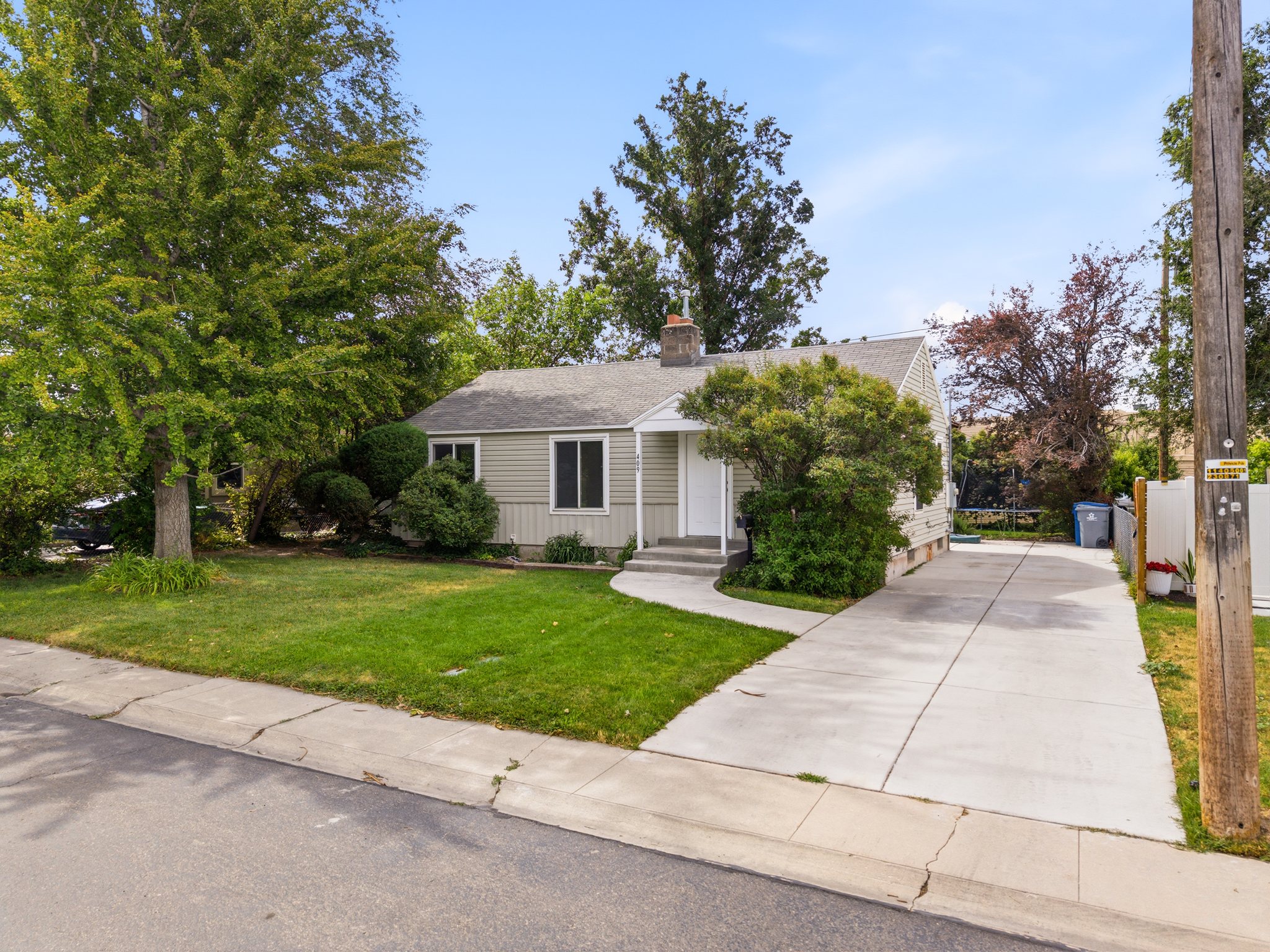 Light green siding exterior with a dark gray shingle roof, mature trees, a manicured lawn, and a concrete driveway