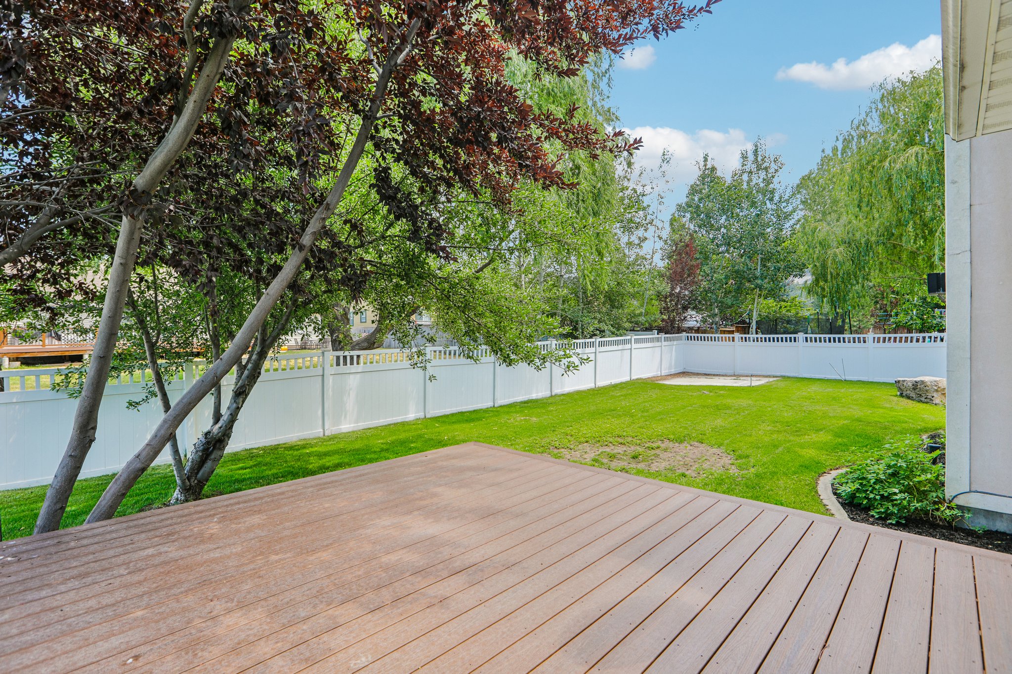 Spacious backyard featuring a composite deck, green lawn, white vinyl fencing, mature trees, and a garden bed