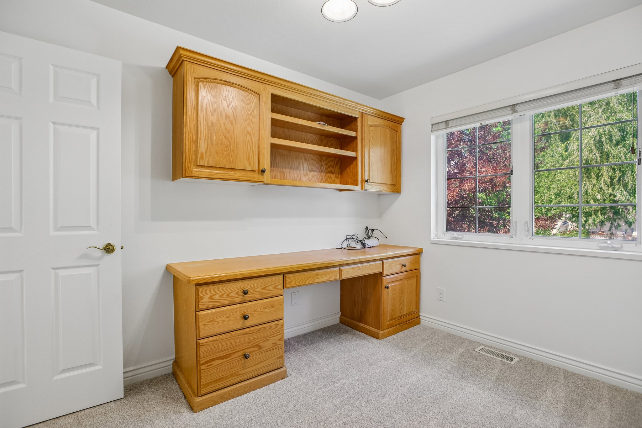 Built-in wood-finish desk with drawers and upper cabinetry, complemented by a large window with exterior foliage