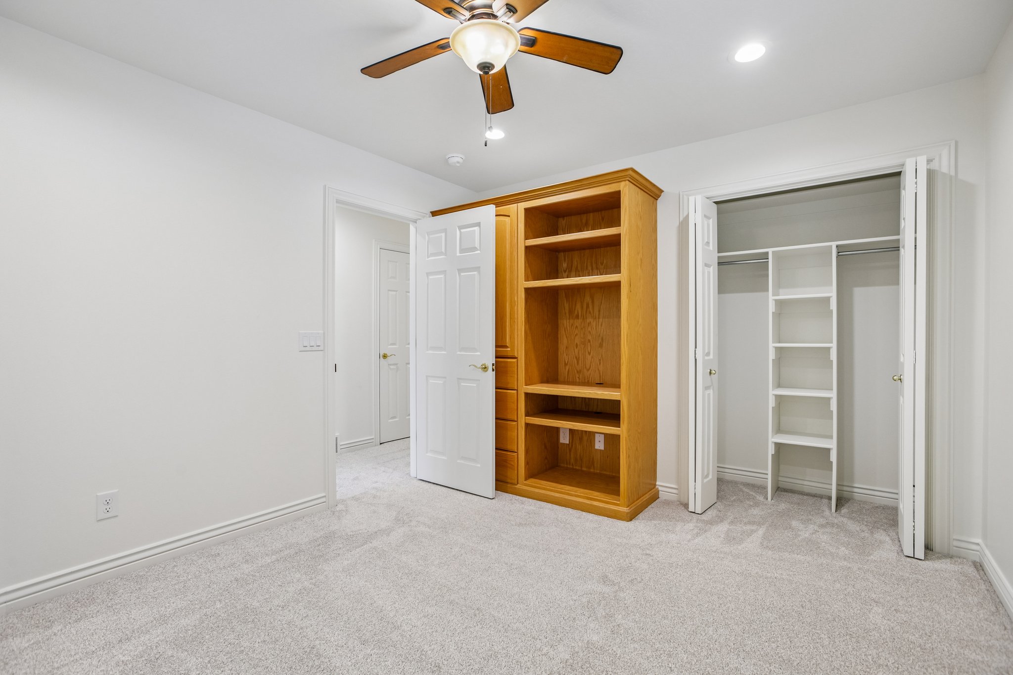Bedroom featuring light gray carpet, white painted walls, a built-in wooden shelving unit, and a ceiling fan with integrated lighting