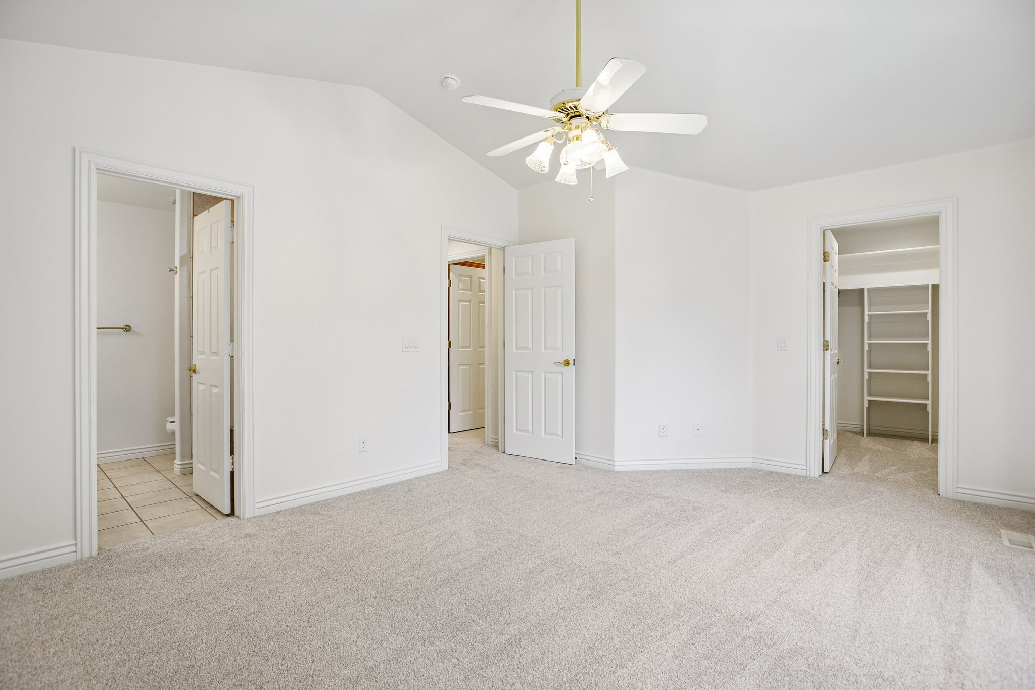 Spacious room featuring vaulted ceilings, light-colored carpeting, a ceiling fan with integrated lighting, and multiple doorways, including one revealing a built-in closet system