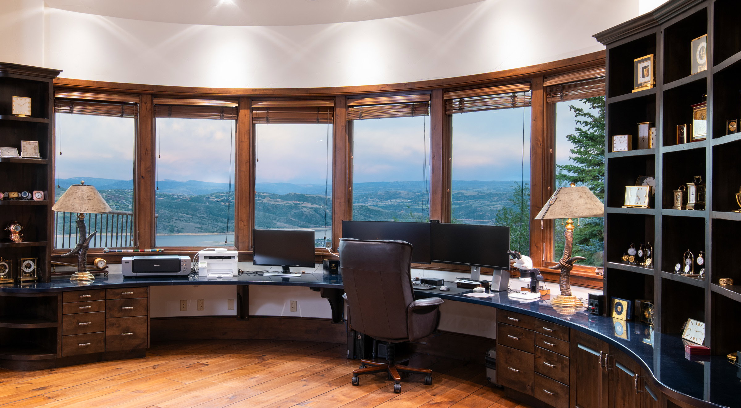 Dedicated office space featuring expansive curved windows, custom built-in cabinetry, integrated shelving, wood-finish flooring, and a natural landscape backdrop