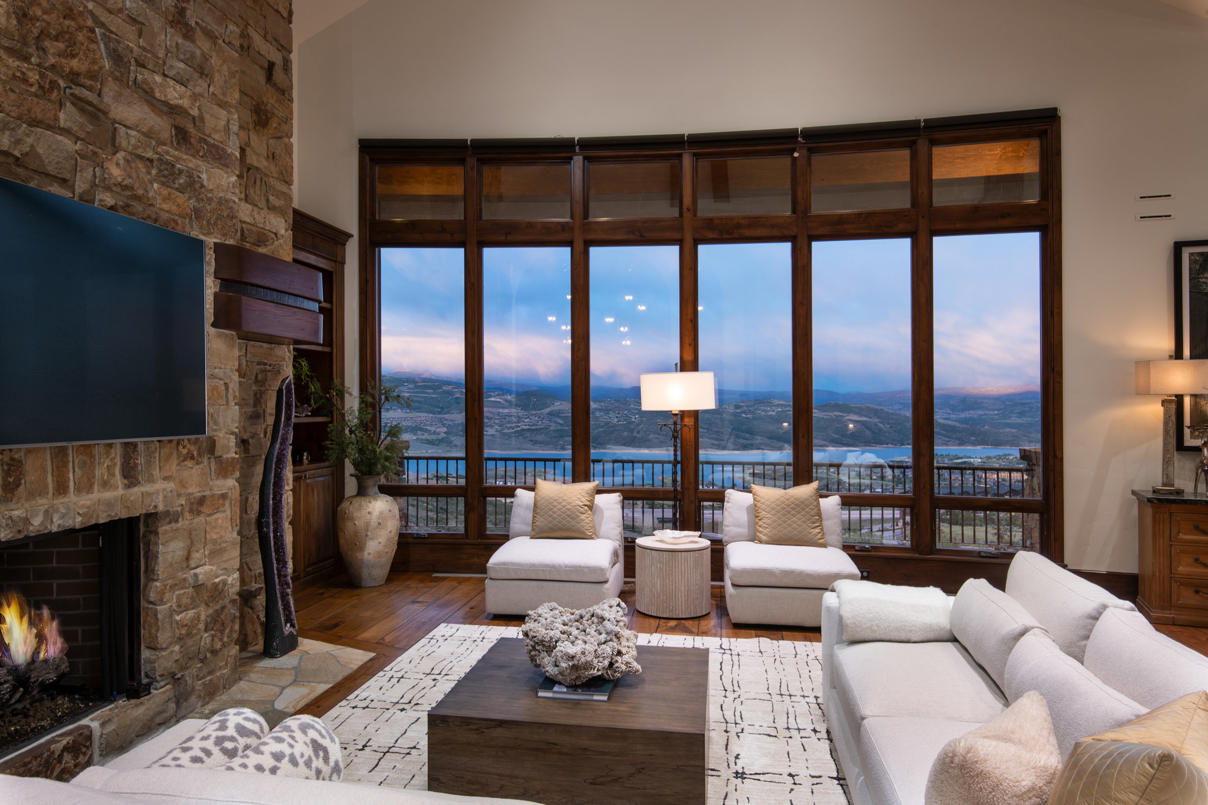 Stone-clad fireplace with hearth, wood-finish flooring, and expansive windows showcasing a distant body of water and natural scenery