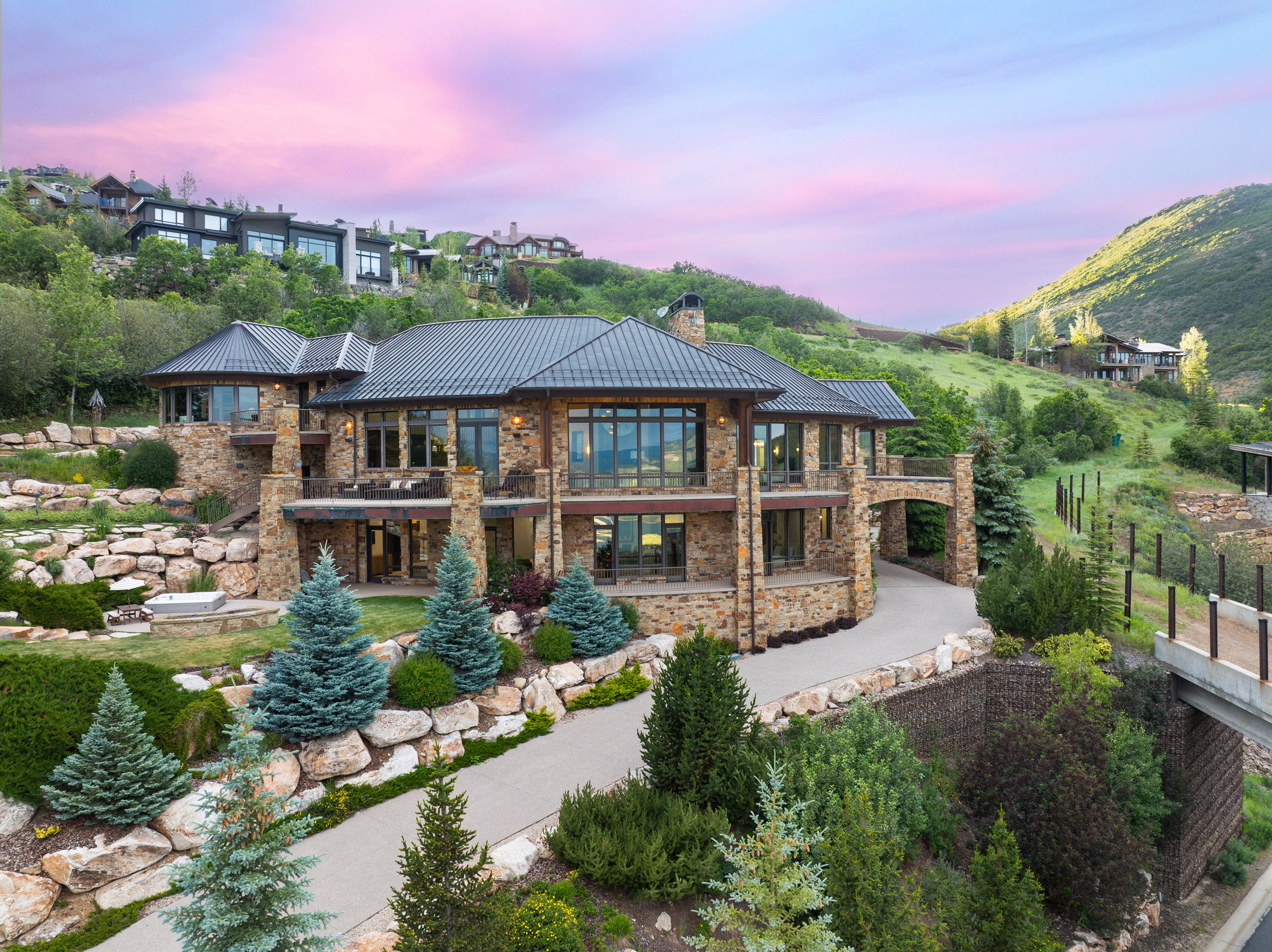 Stone-clad estate featuring multiple balconies, extensive landscaping, a curved driveway, and a prominent arched porte-cochère