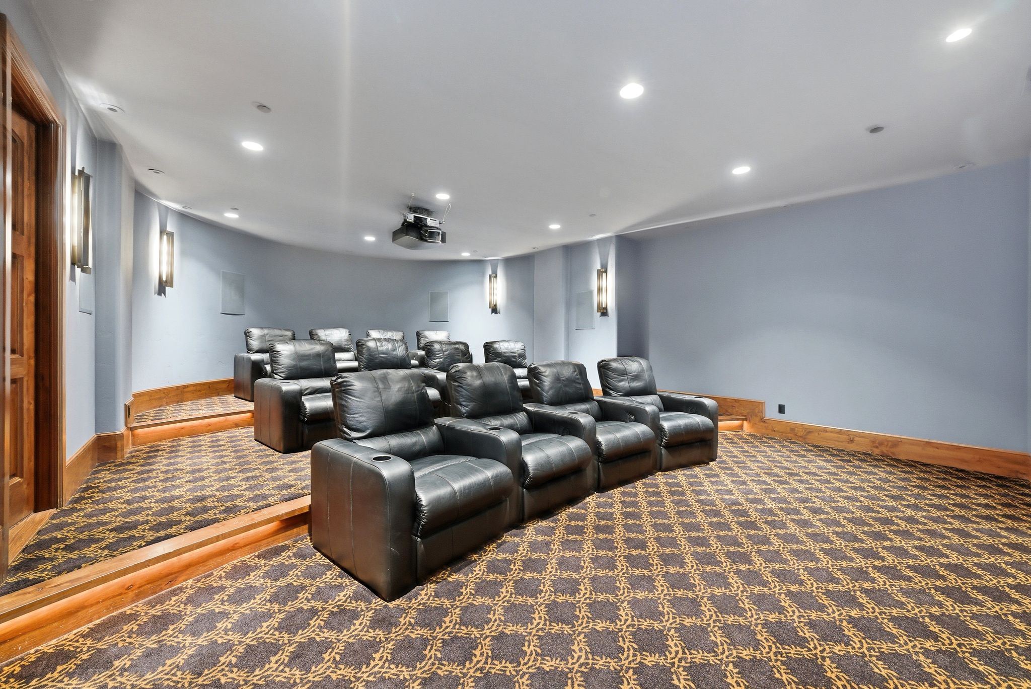 Dedicated theater room featuring tiered seating, patterned carpet, recessed lighting, wall sconces, and a projector