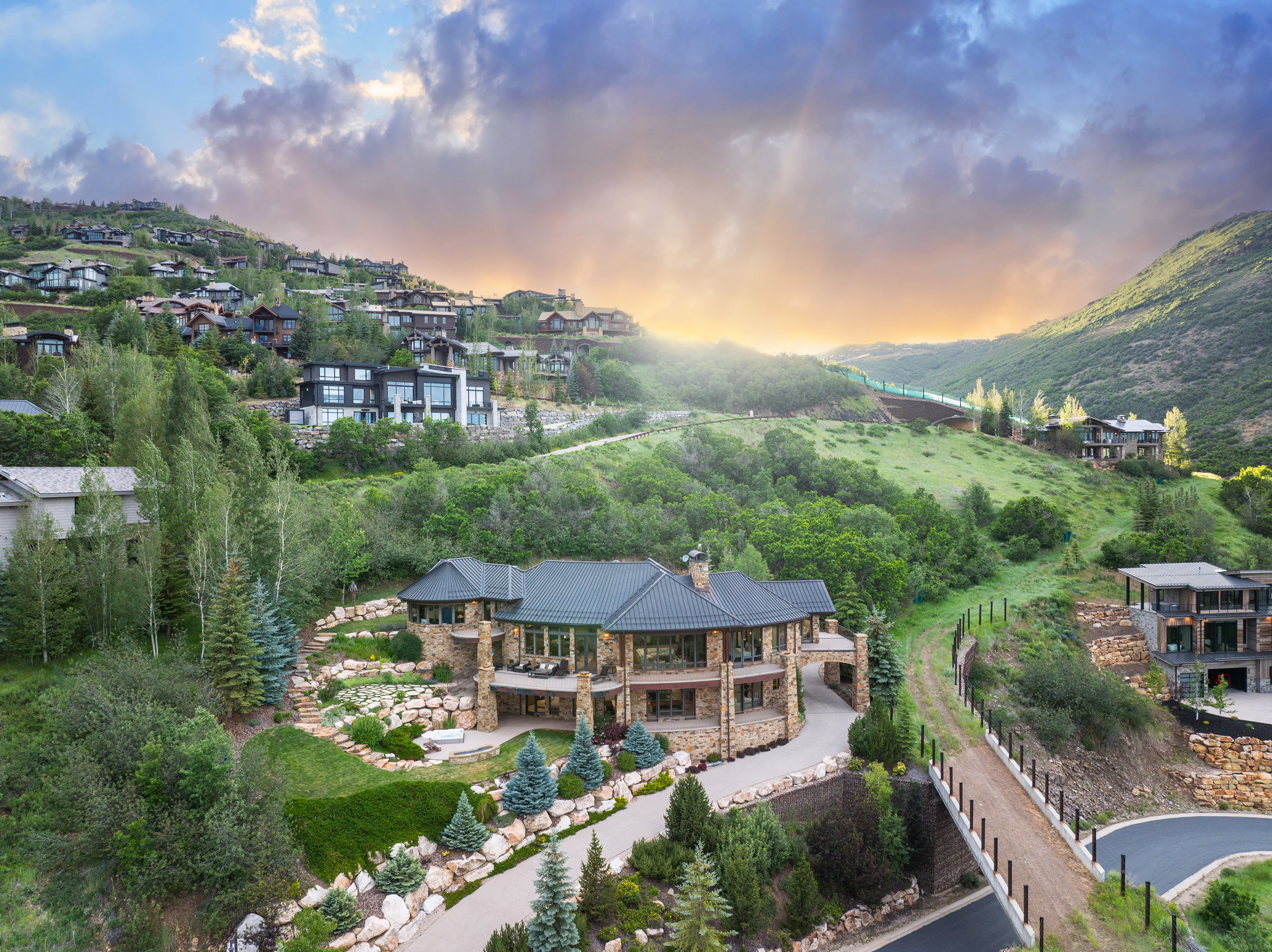 Stone-clad residence featuring multiple balconies, terraced landscaping, a prominent driveway, and a natural mountain backdrop