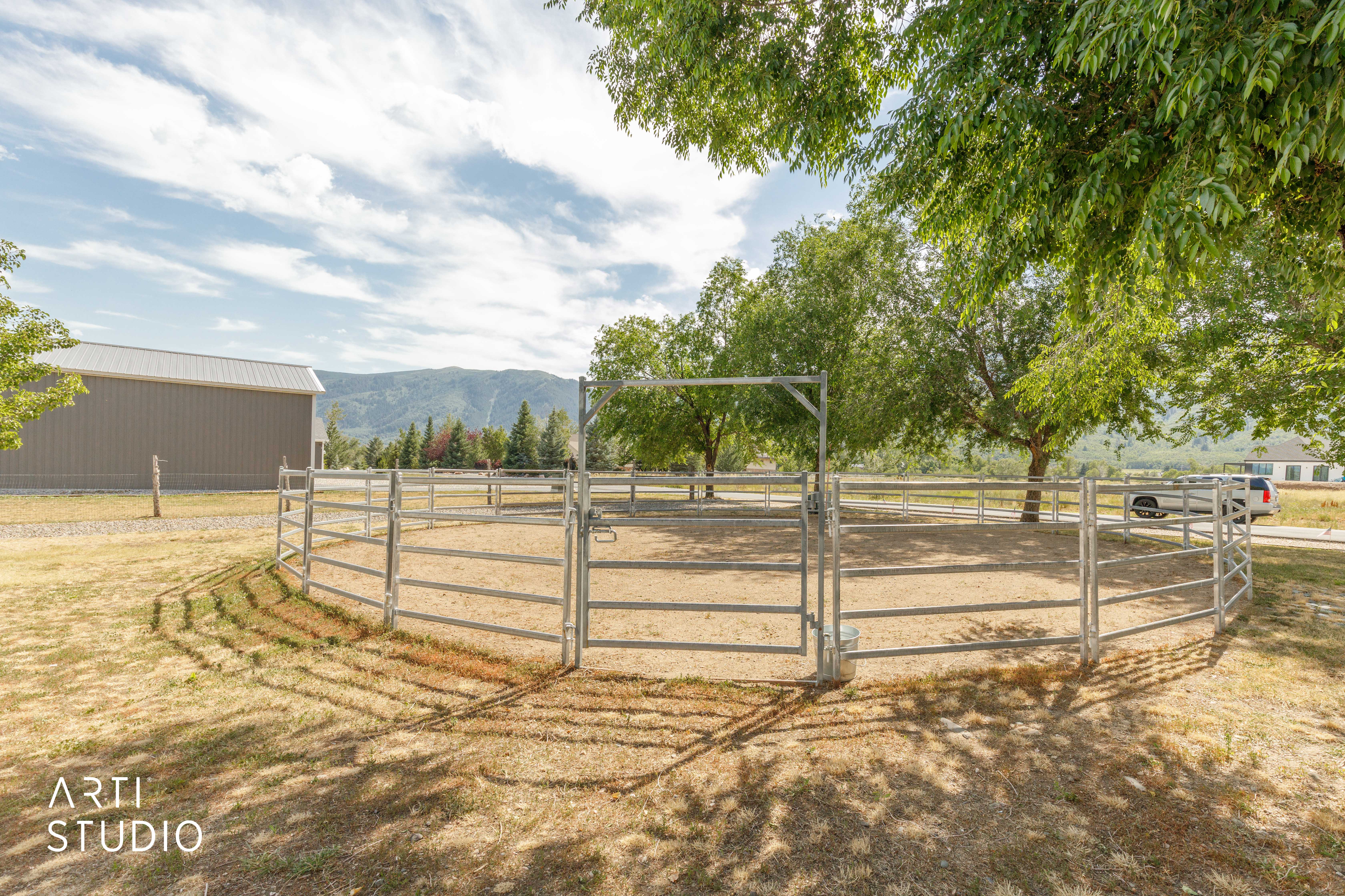 Circular metal fenced enclosure with a gravel base, bordered by mature deciduous trees and a distant mountain range