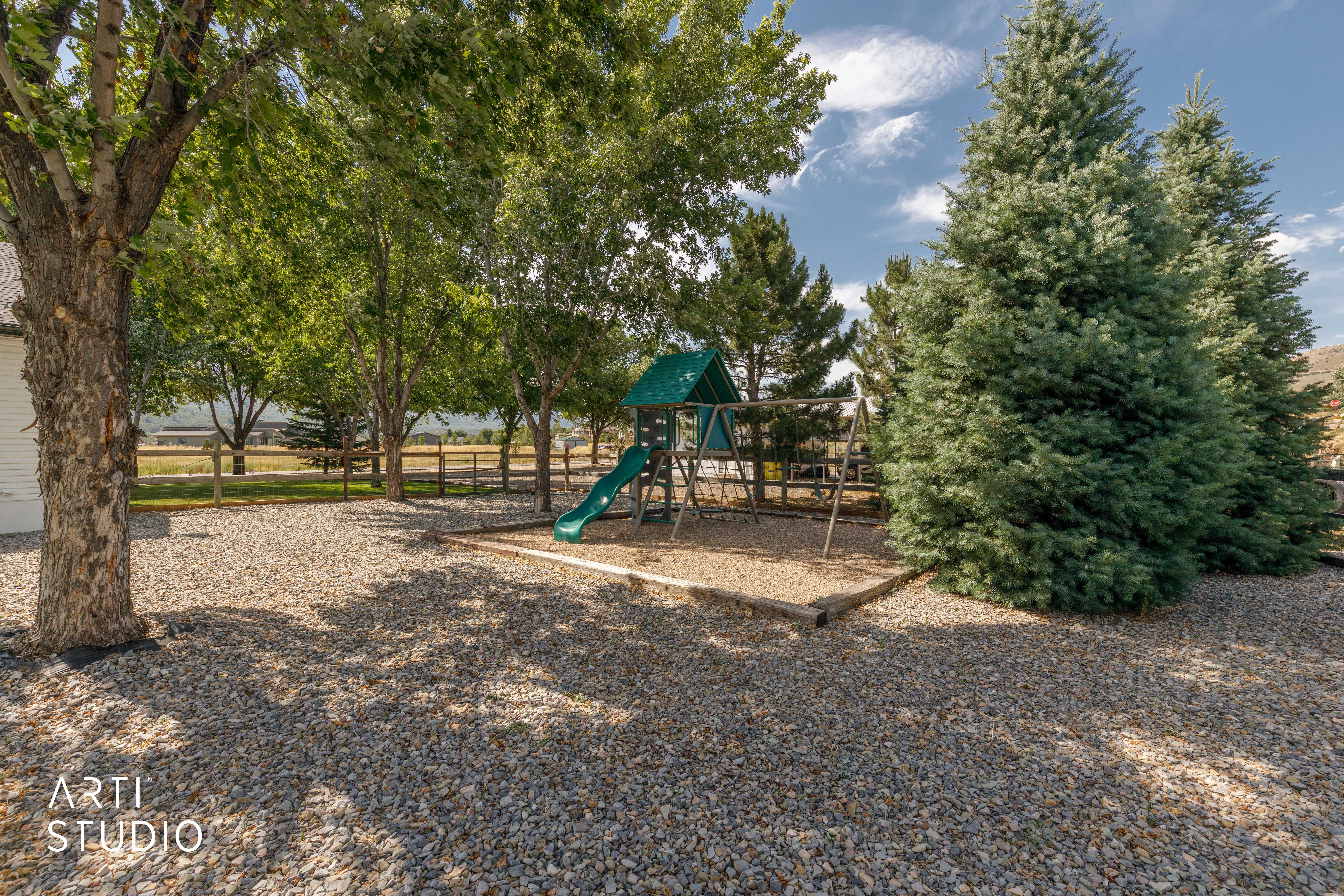 Gravel-covered outdoor recreation area featuring mature trees, a wooden fence, and a playground structure with a green slide