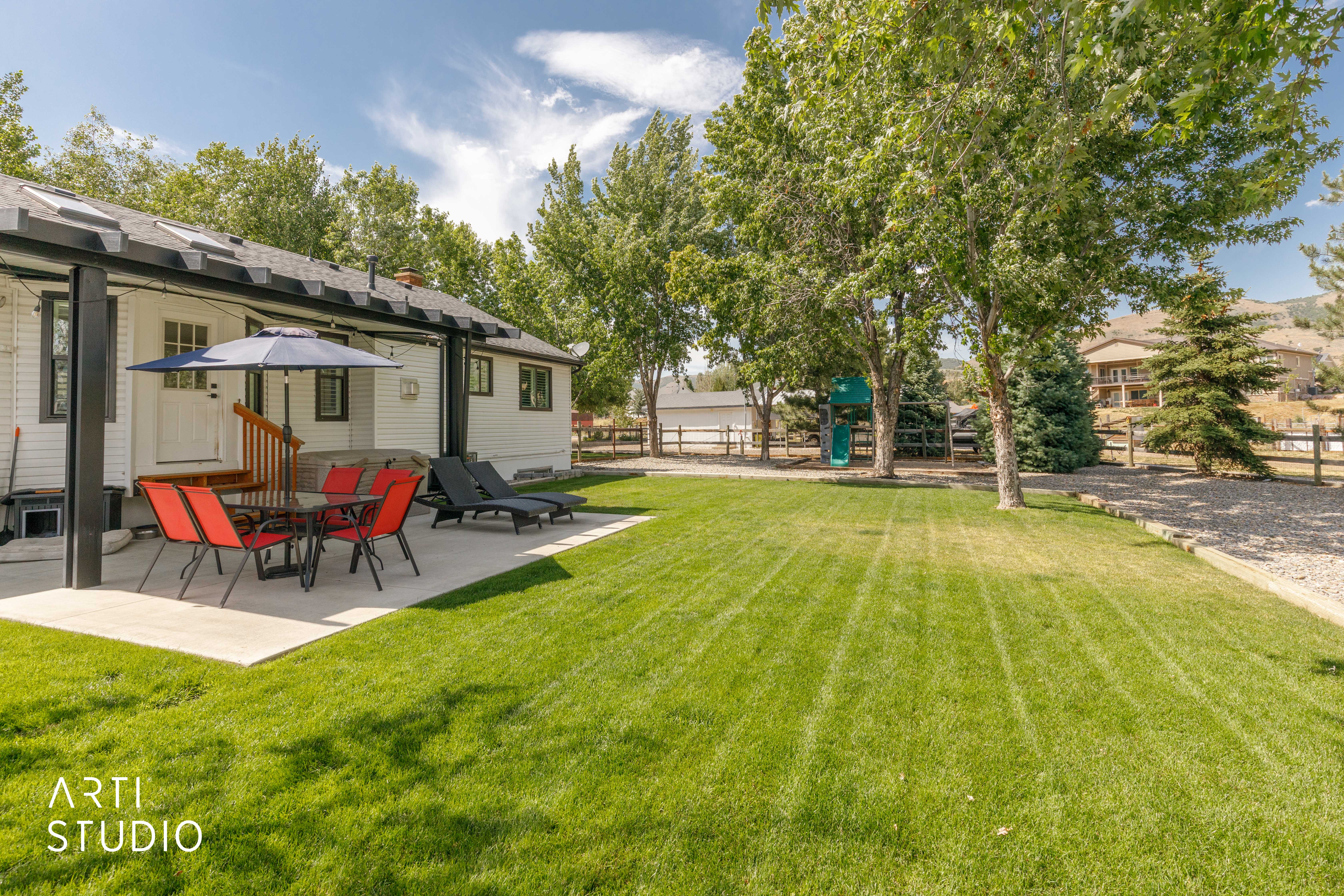 Expansive backyard featuring a large concrete patio with a covered pergola, a lush green lawn, mature trees, and a privacy fence