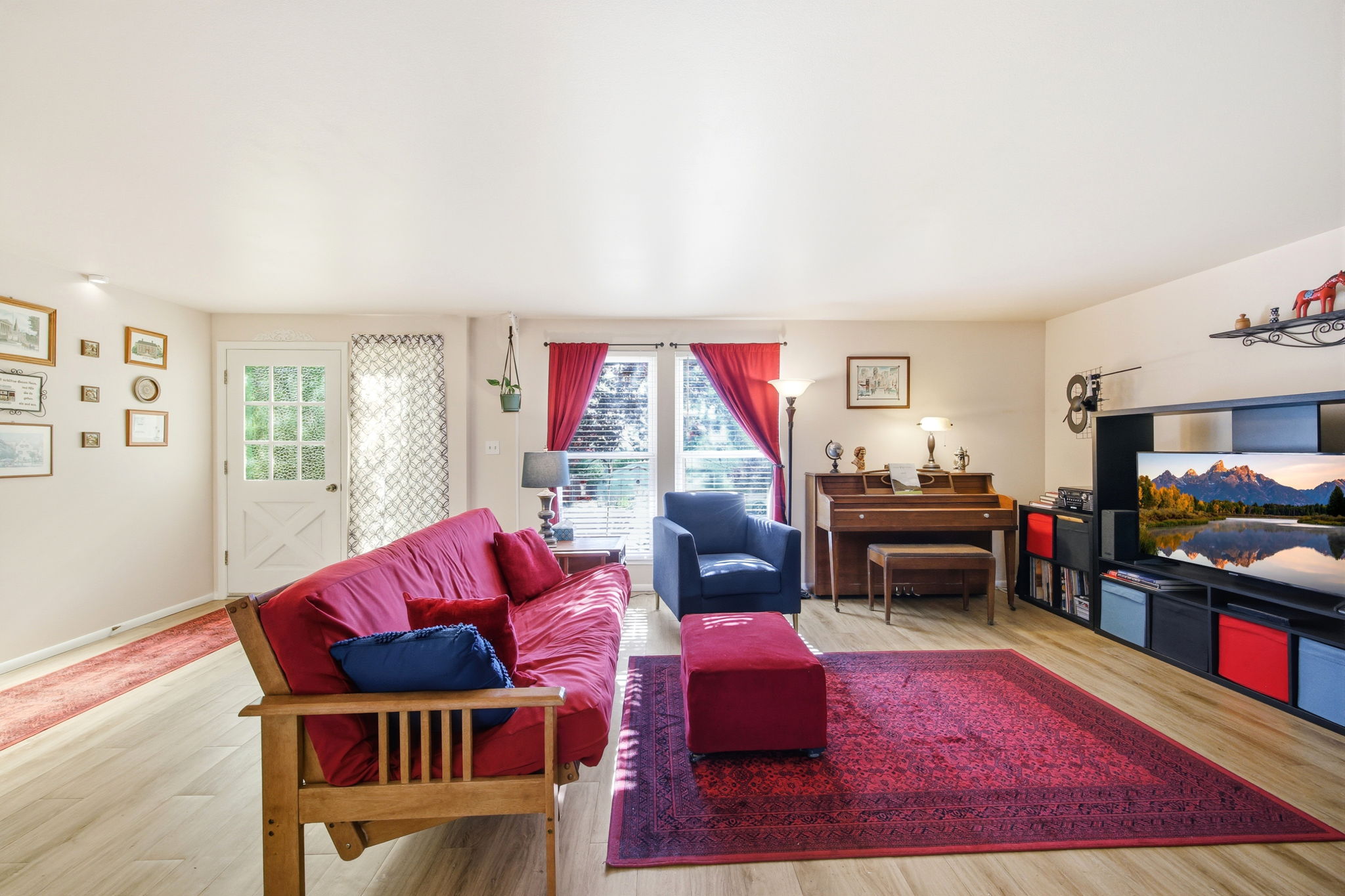 Spacious living area featuring light wood-finish flooring, a white glass-paneled door, and large windows with red drapes providing natural illumination