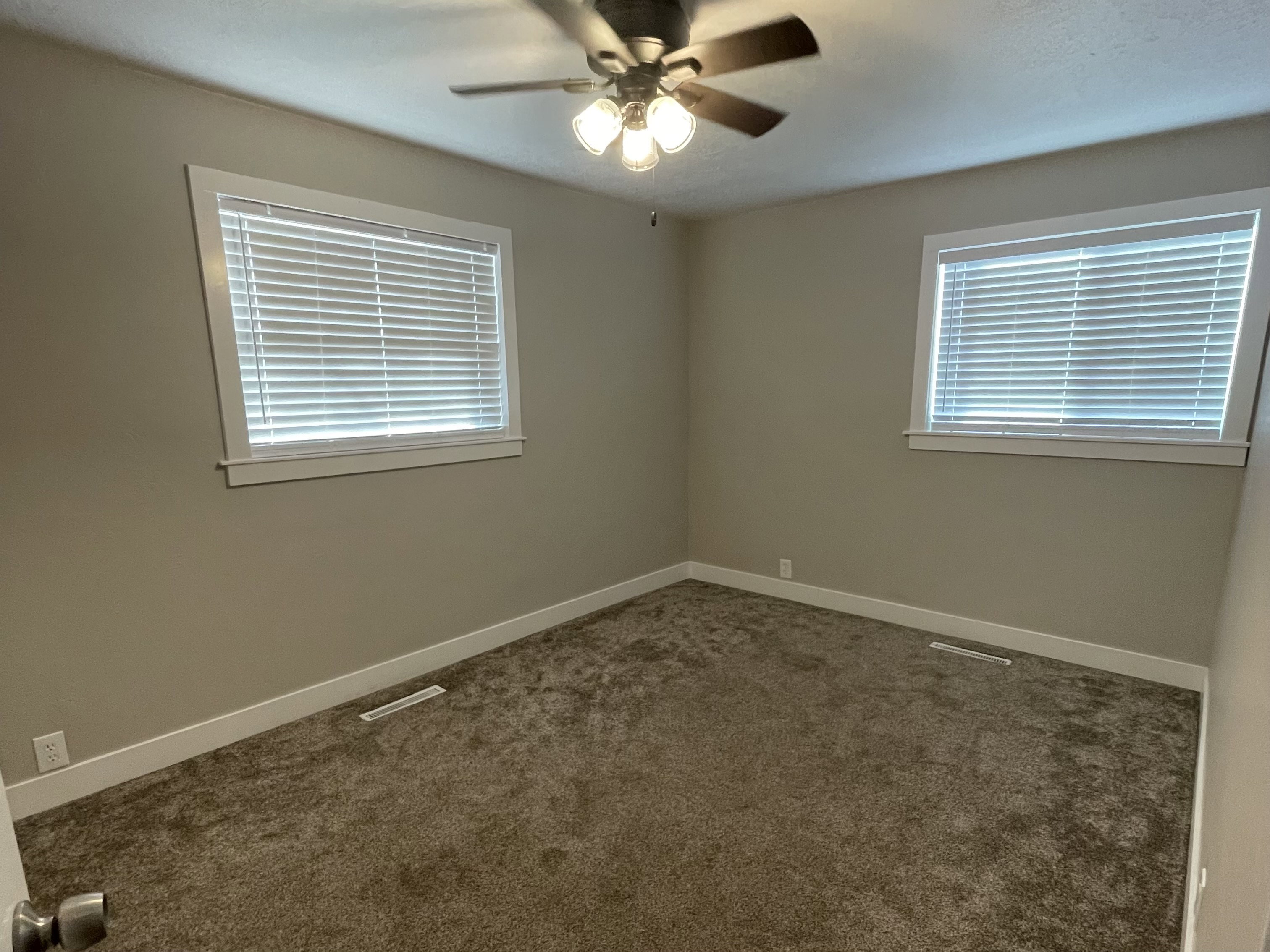 Carpeted room featuring a ceiling fan with light fixture, two windows with blinds, neutral wall paint, and white baseboards