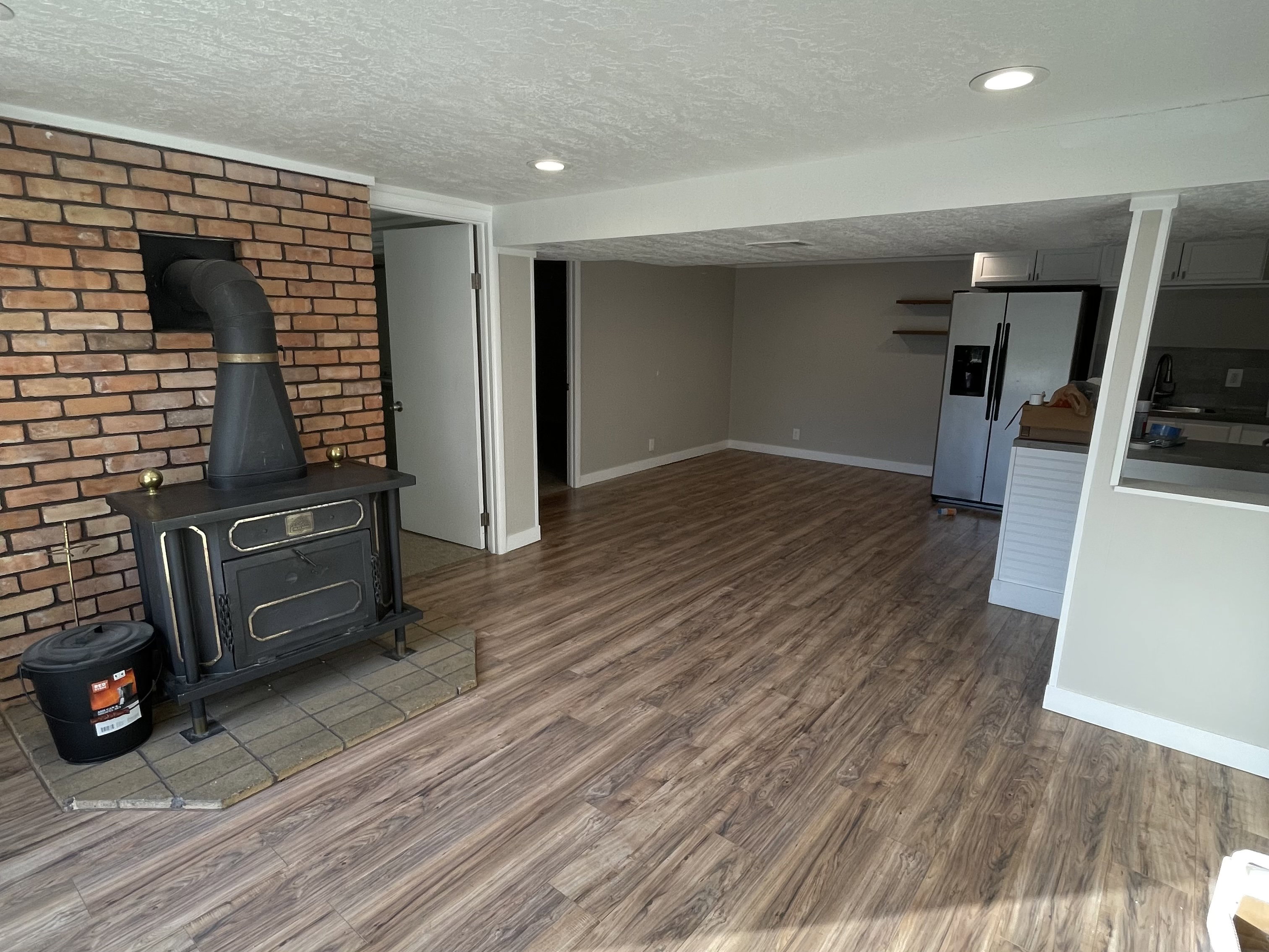 Open-concept living area featuring wood-finish flooring, recessed lighting, and a brick accent wall with a wood-burning stove