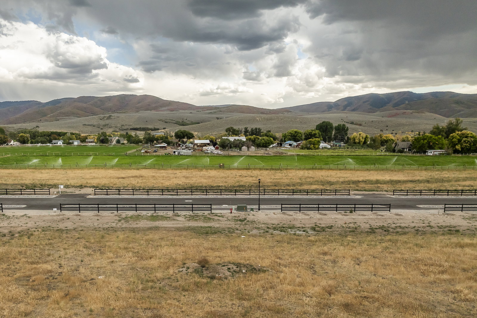 Expansive property featuring a paved road, dark fencing, agricultural land with active sprinklers, and distant residential structures against a backdrop of rolling hills