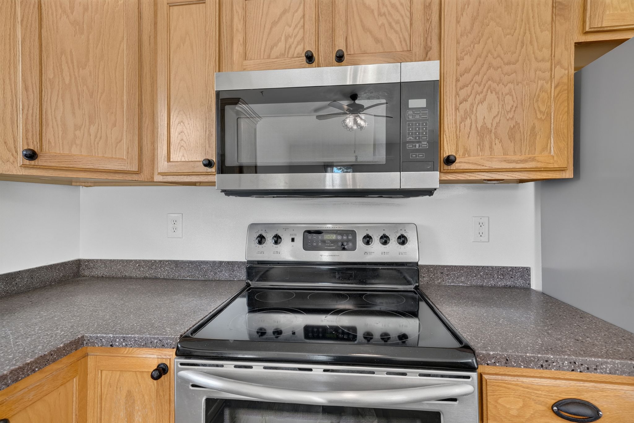 Kitchen featuring a stainless steel oven with a smooth cooktop, a coordinating stainless steel microwave, light wood cabinetry with dark hardware, and speckled gray countertops