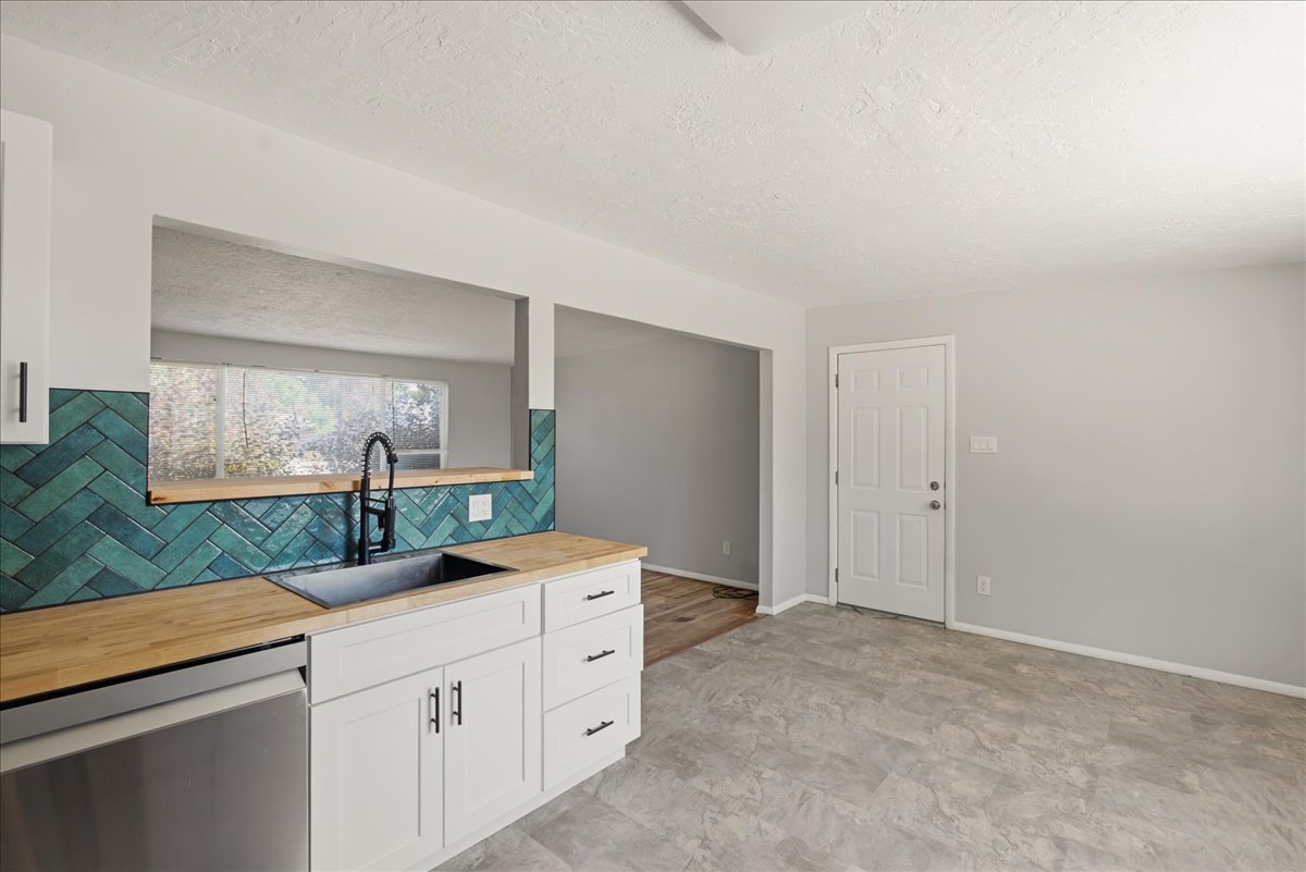 Kitchen featuring white cabinetry with dark hardware, butcher block countertops, a black sink with a black pull-down faucet, a teal herringbone pattern tile backsplash, and stainless steel dishwasher
