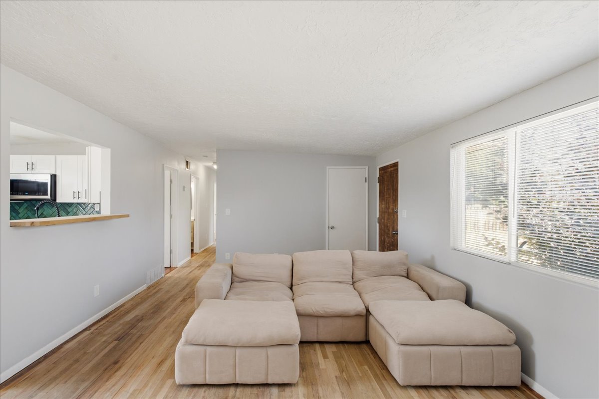 Living area featuring wood-finish flooring, a pass-through window to the kitchen, and a large window with blinds