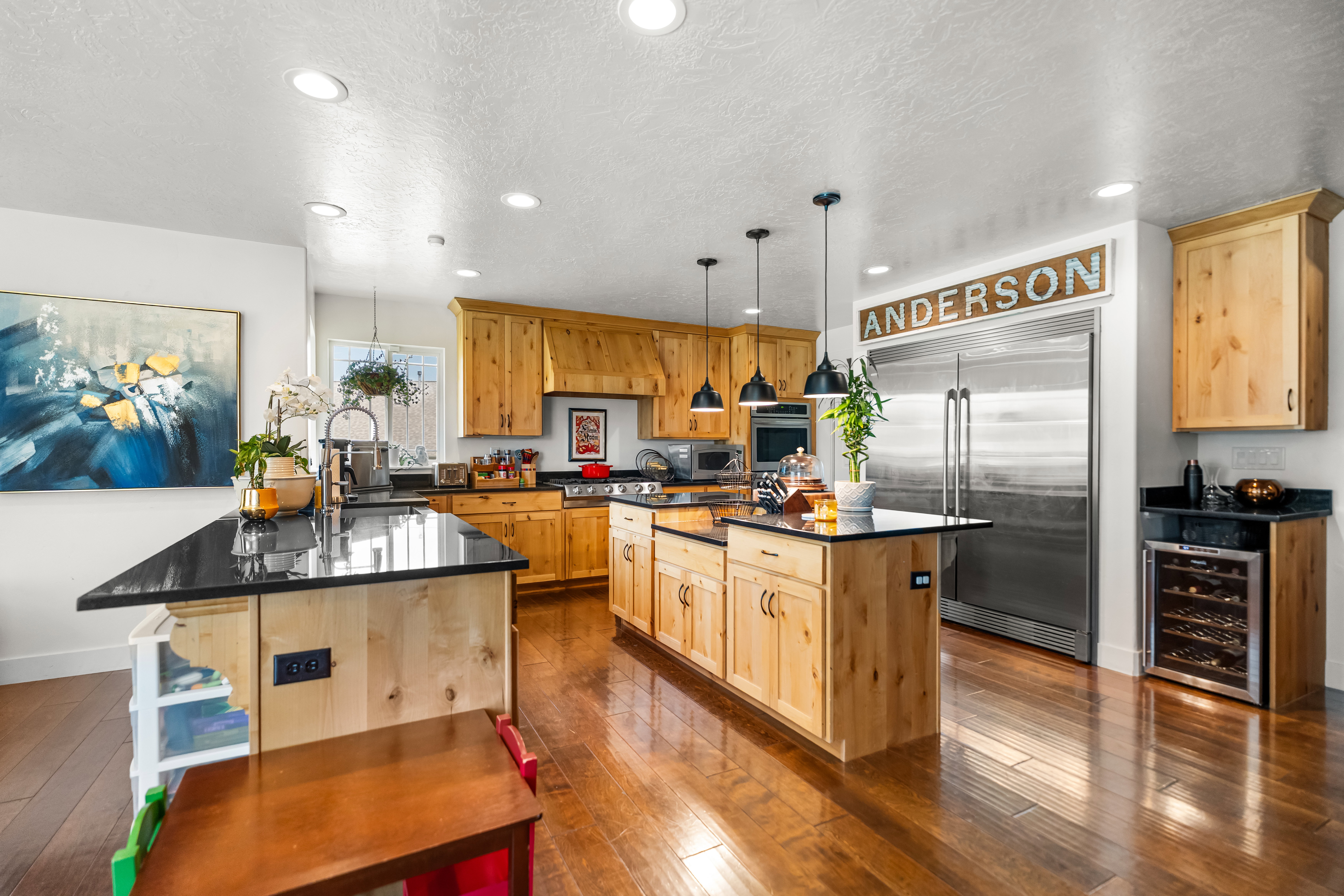 Spacious kitchen featuring natural wood cabinetry, dark countertops, stainless steel appliances, a large central island with pendant lighting, and wood-finish flooring
