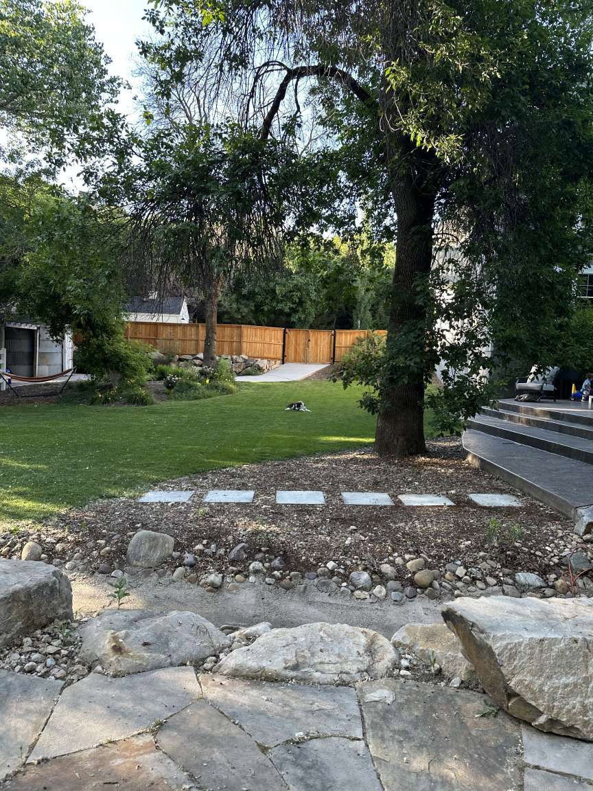 Expansive backyard featuring a natural stone patio, a large grass lawn, mature trees, a wood privacy fence, and concrete stepping stones
