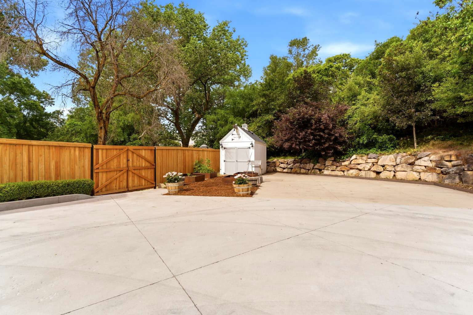 Spacious concrete pad featuring a privacy fence with double gates, a white storage shed, and a natural stone retaining wall