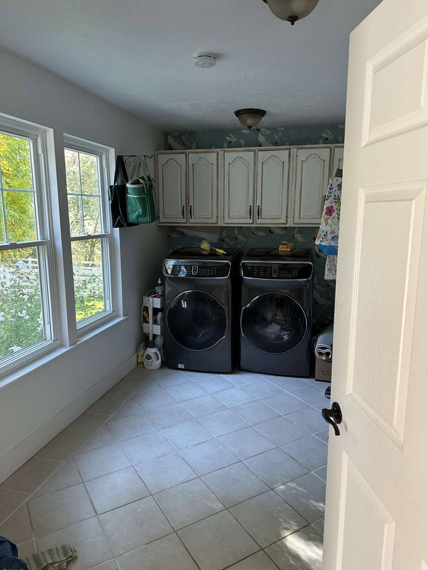 Laundry space featuring light-toned tile flooring, two large windows, white cabinetry with dark hardware, and a ceiling-mounted light fixture
