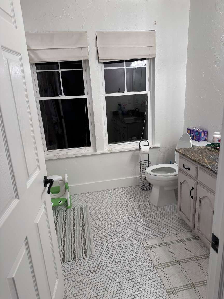 Bathroom featuring white hexagonal tile flooring, dual windows with Roman shades, a vanity with a stone countertop, and a white paneled door with black hardware