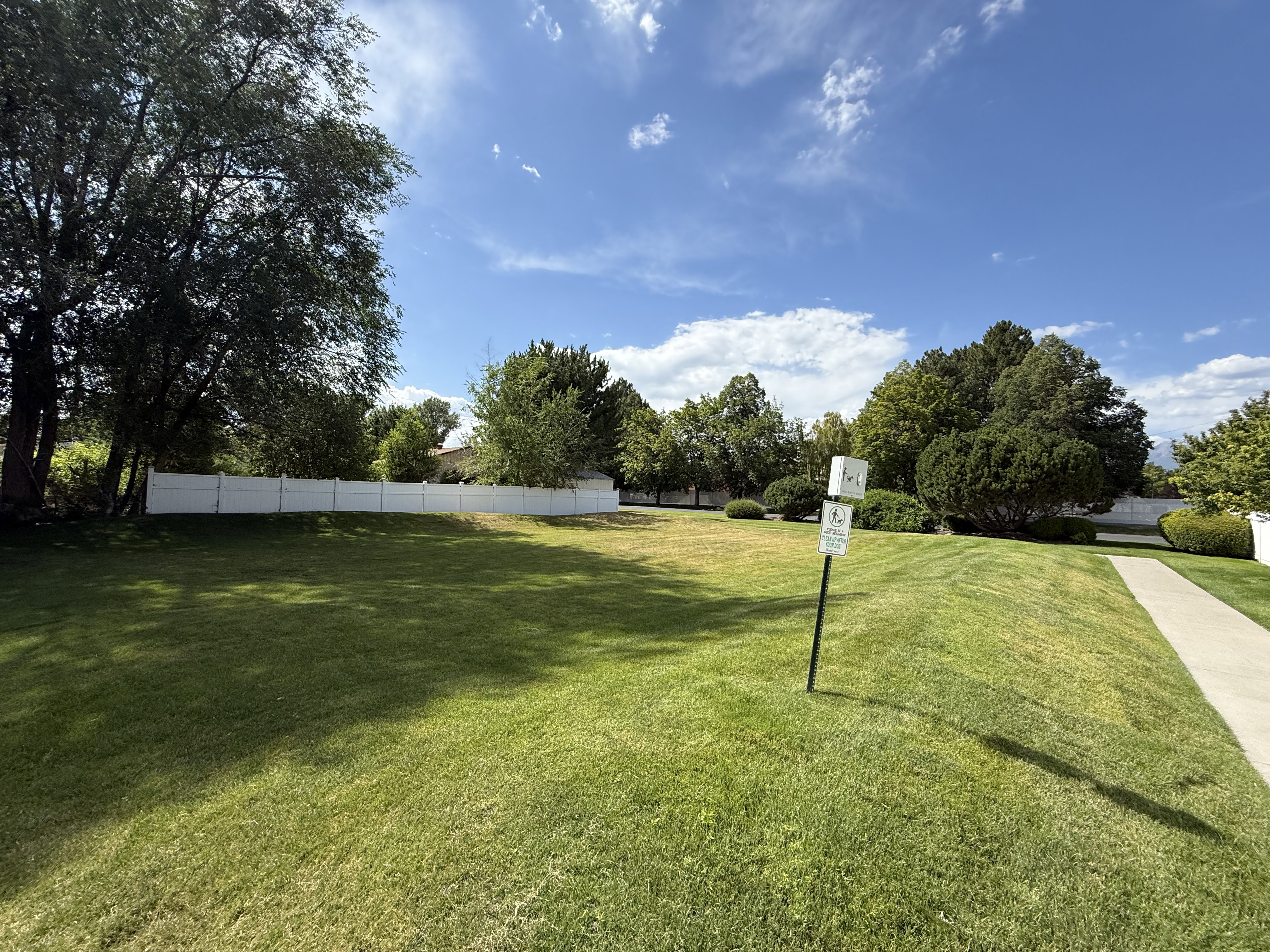 Expansive lawn area featuring a white privacy fence, mature trees, a paved walkway, and maintained landscaping