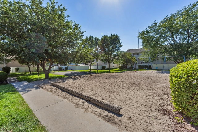 Expansive sand play area featuring a large log element, adjacent to a concrete pathway and mature trees