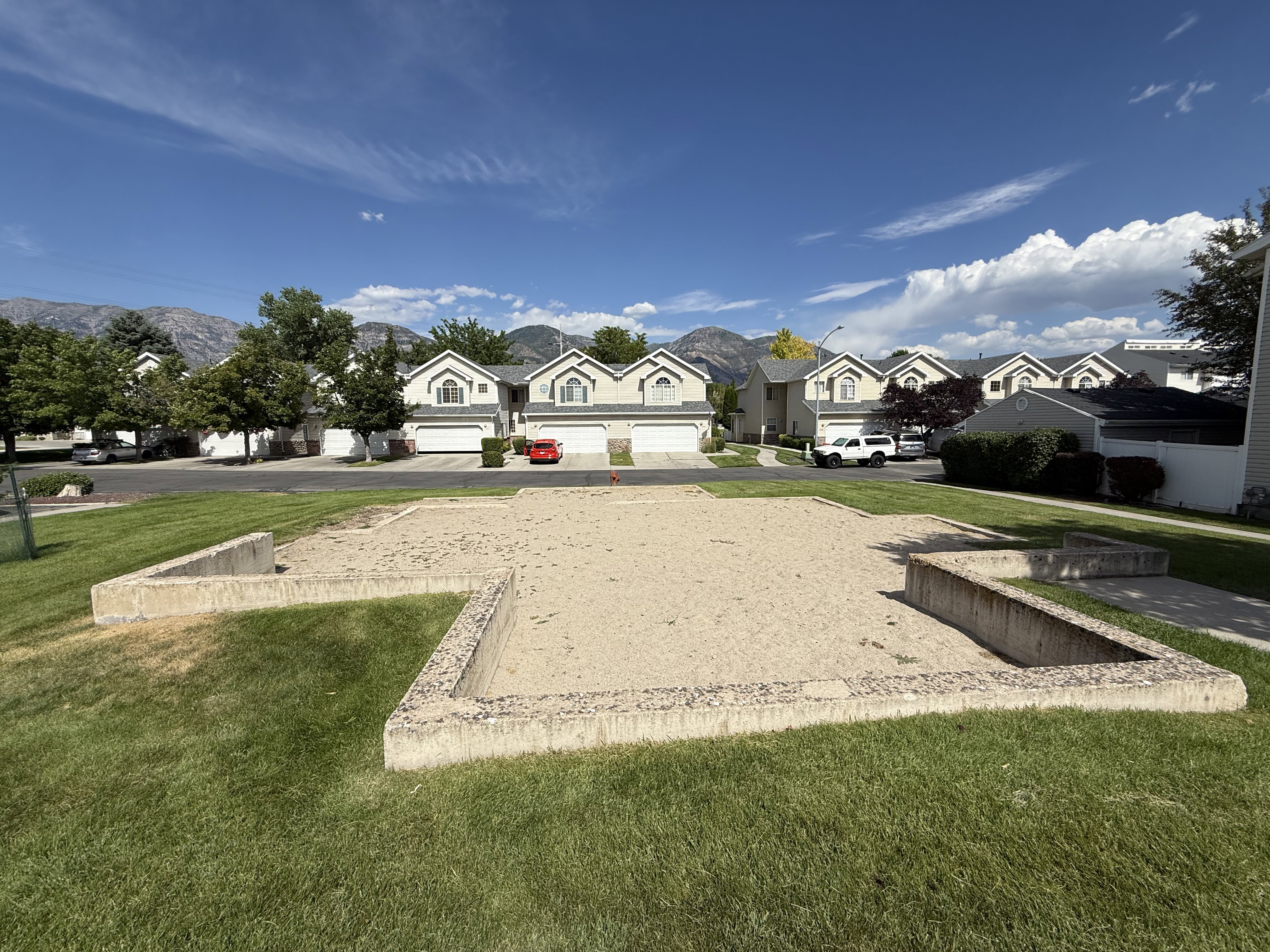 Community green space with a sand pit surrounded by concrete edging, manicured lawn, mature trees, and mountain backdrop